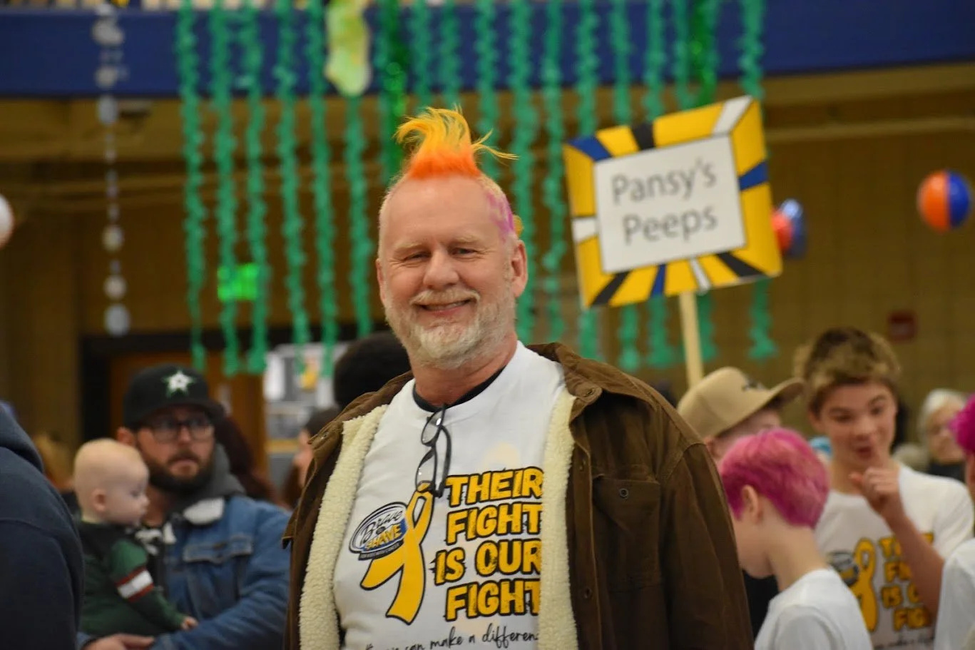 Smiling man with colorful mohawk hairstyle in a crowd at a charity event, wearing a white T-shirt with a yellow ribbon symbol and text. In the background, a sign reads 'Pansy's Peeps'.