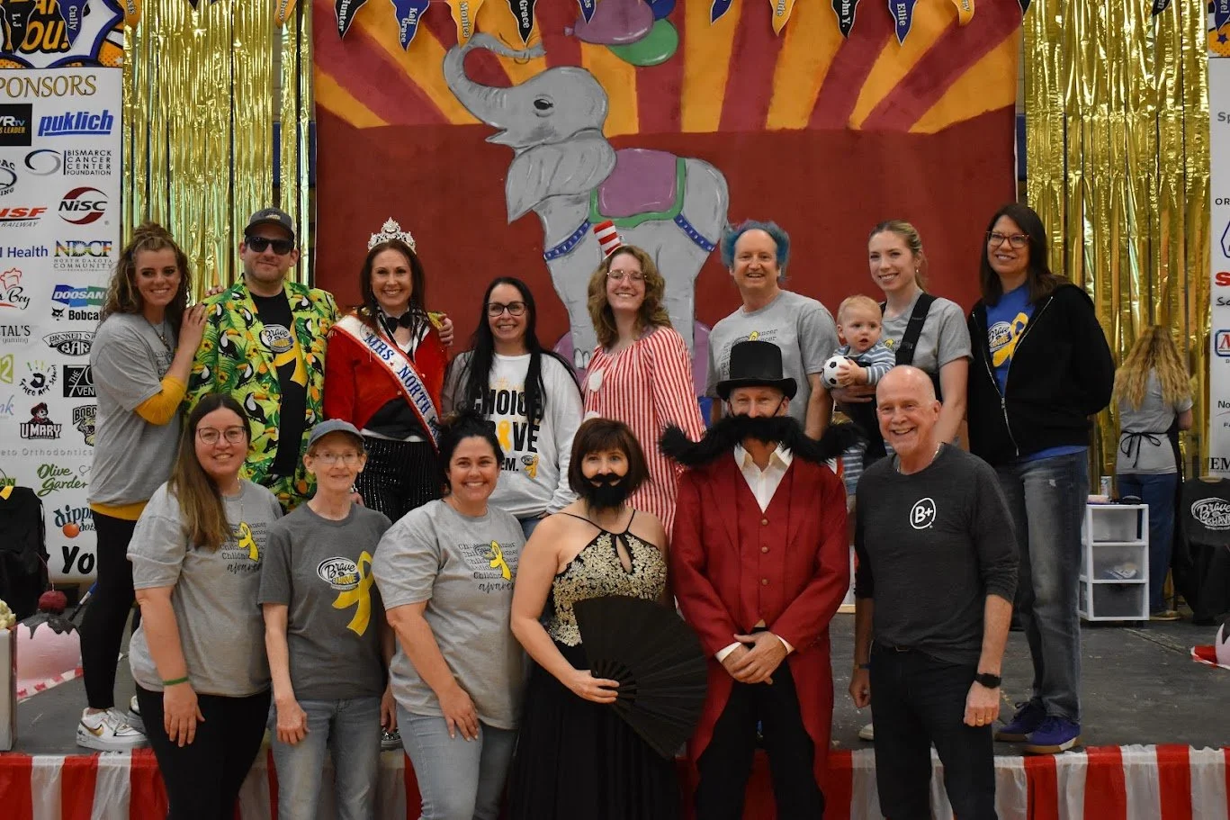 A group of people on a stage, some wearing costumes, including a woman with a crown and sash, and a man with a top hat and red coat. The backdrop has a cartoon elephant, and the stage is decorated with gold streamers.