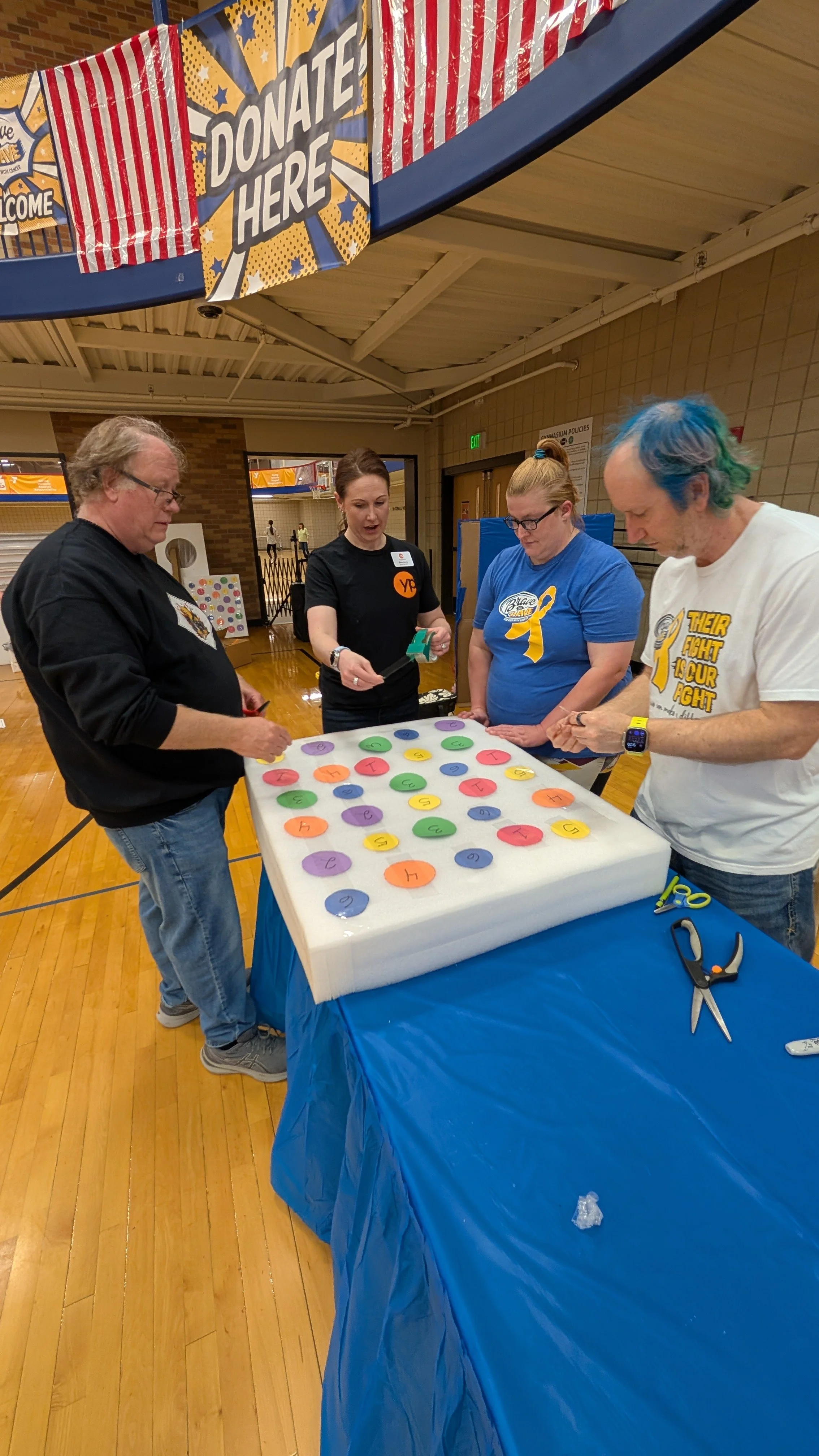 Four people stand around a table with colored circles on it, inside a gymnasium. They are participating in a donation event, with a patriotic banner overhead that says 'DONATE HERE.' The table is covered with a blue tablecloth, and there are scissors and other supplies on the side.