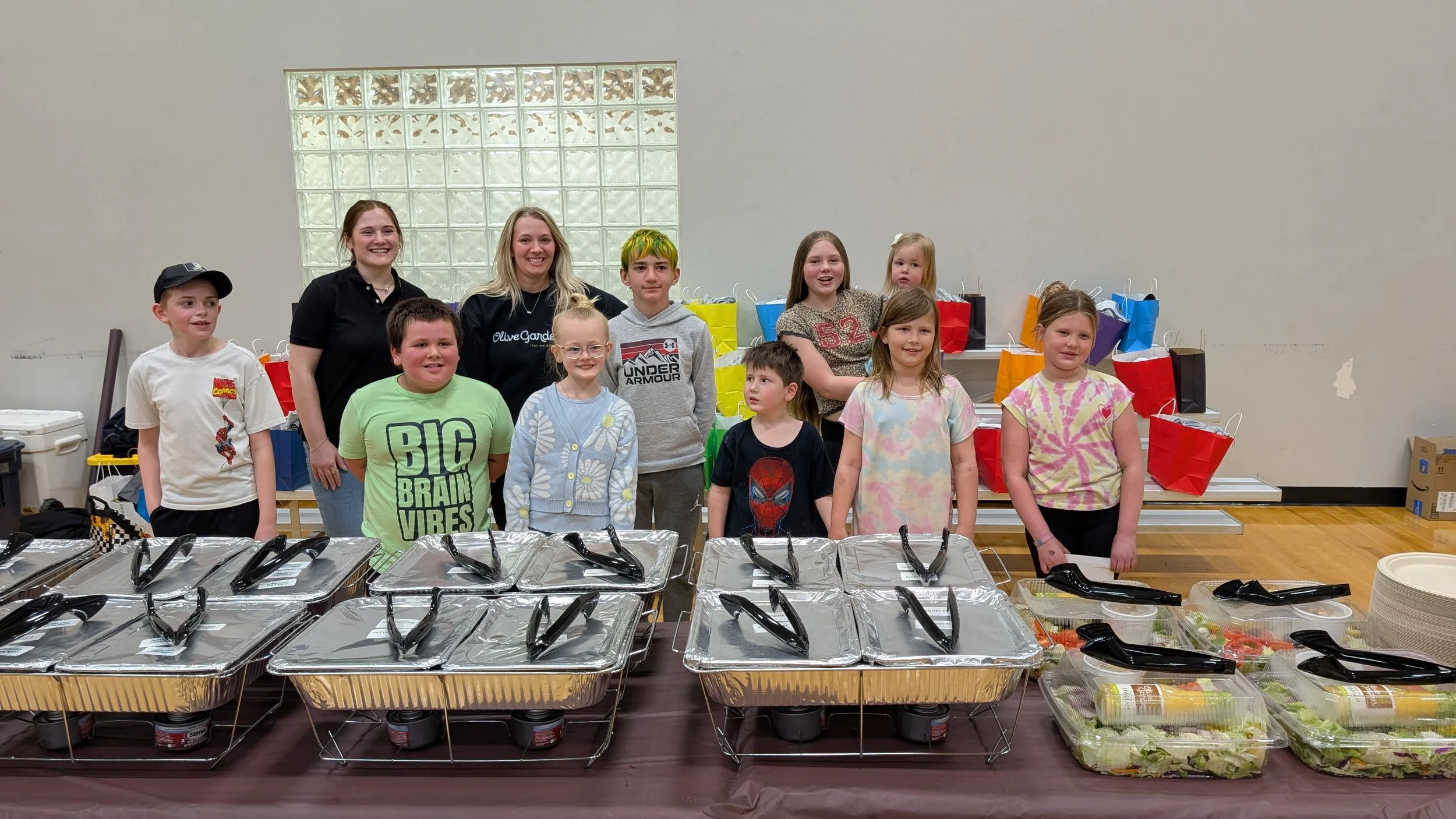 Group of children and two women smiling indoors around a table with food and catering trays, with gift bags in the background.
