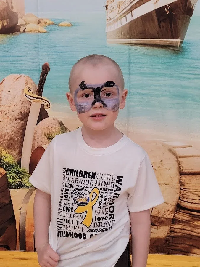 A young boy with face paint resembling a butterfly, standing in front of a beach-themed backdrop with rocks, water, and a ship. He is wearing a white T-shirt with words related to childhood cancer awareness.