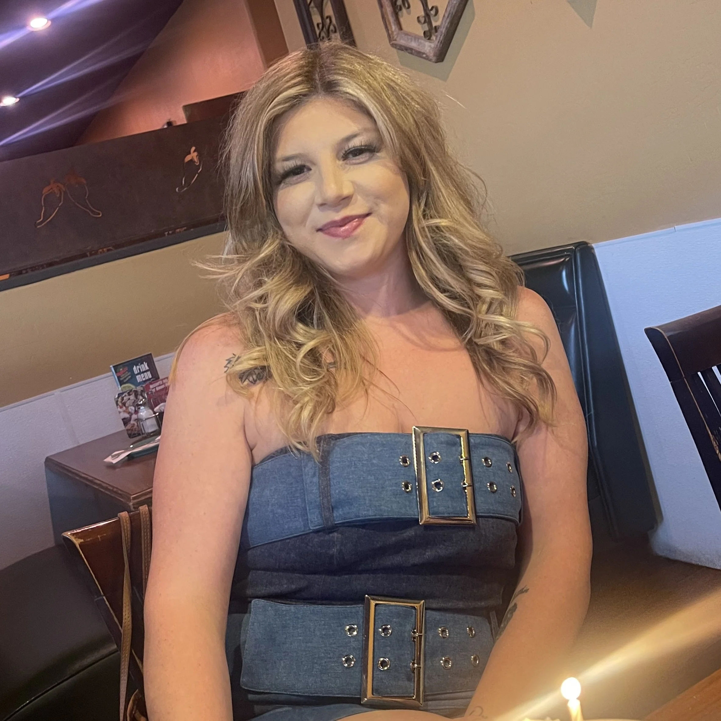 A woman with long, curly blonde hair wearing a dark strapless top with metallic embellishments, smiling at the camera inside a restaurant or cafe. In the background, there is a wall with framed artwork and a menu stand on the table.