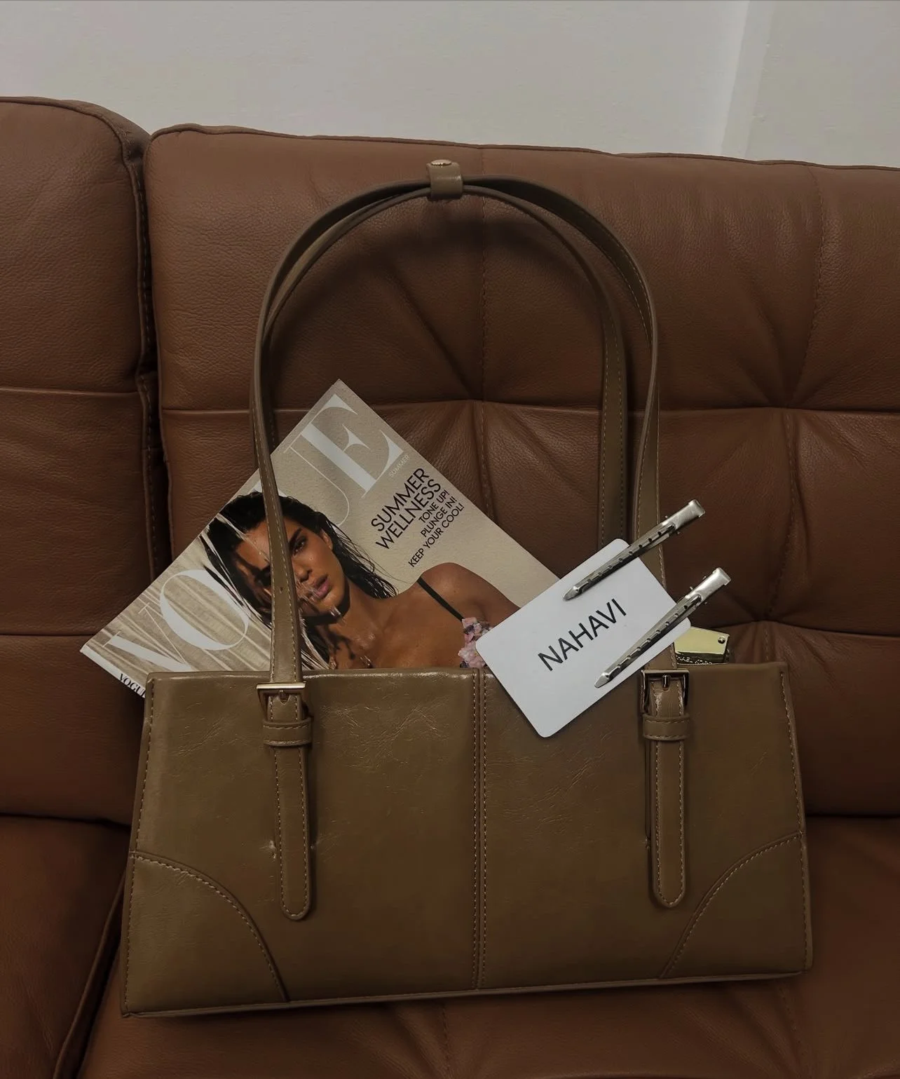 A beige handbag with two metal clips holding a white card that says "NAHAV". Inside the bag, there is a magazine featuring a woman with dreadlocks, and a folded paper with the word "SUMMER" partially visible. The items are placed on a brown leather couch.