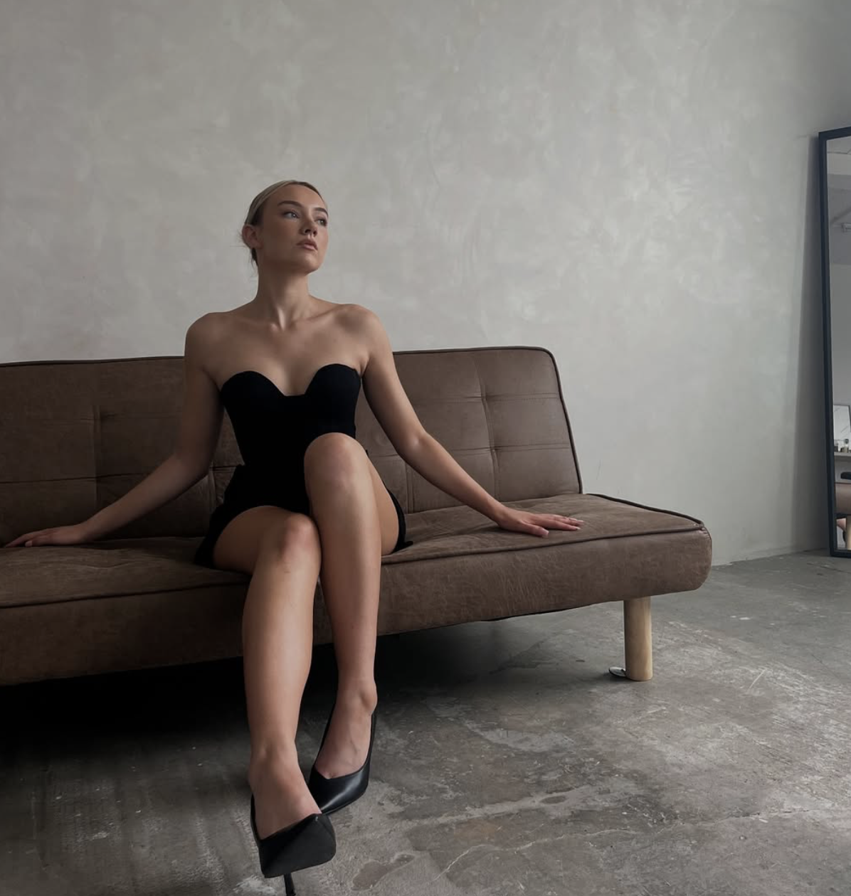 A woman with blonde hair wearing a strapless black dress and black high heels sits on a brown sofa in a minimalist room with a plain white wall. She has a confident pose, with one leg crossed over the other and her hands resting on the sofa.