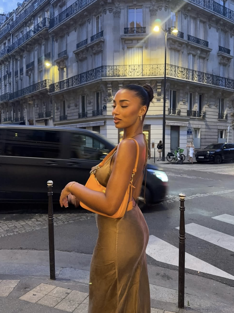 A woman in a beige dress holding an orange bag standing on a city sidewalk at night with a Parisian building in the background. A black car passes by and streetlights illuminate the scene.