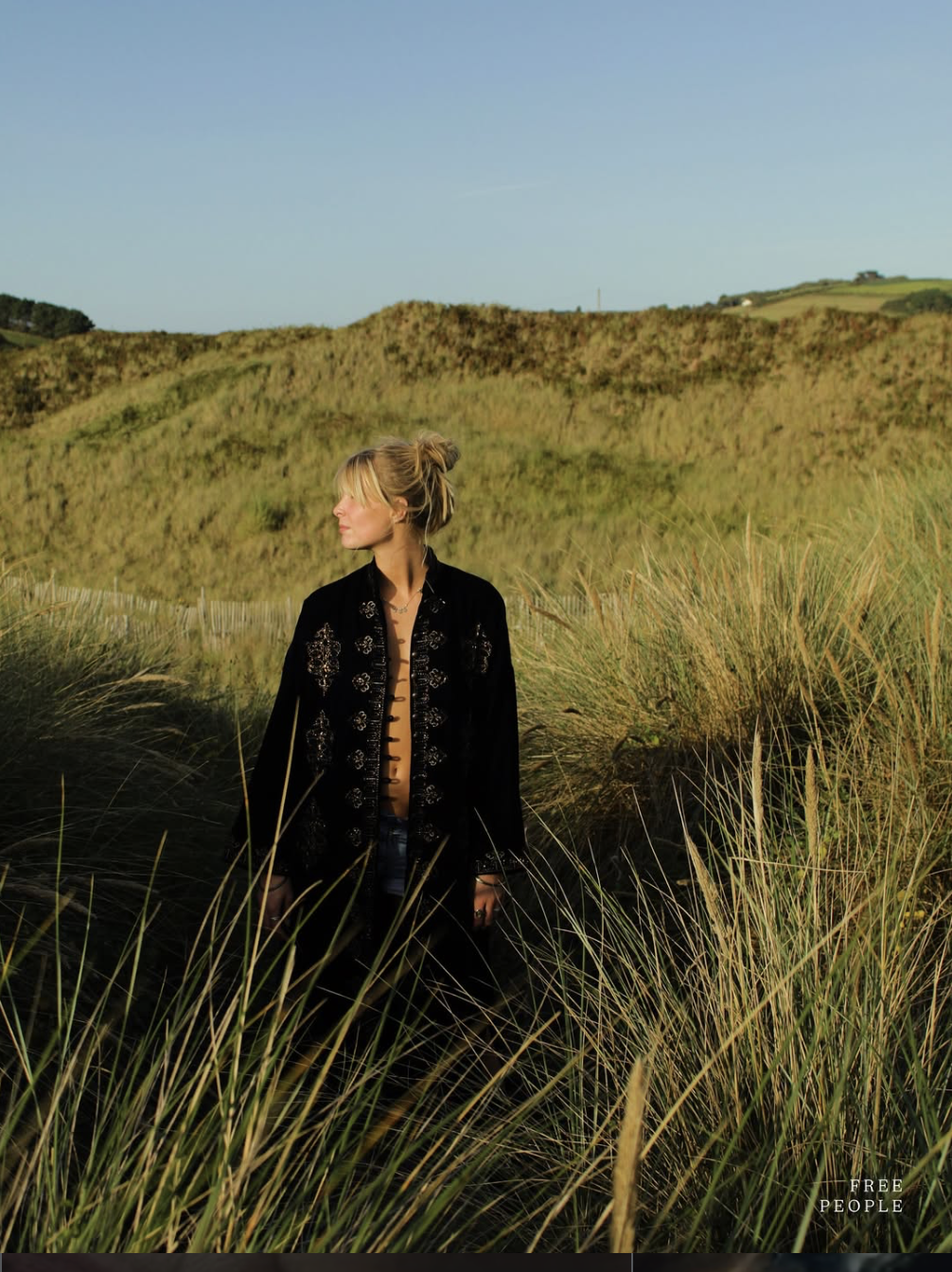 A woman with blonde hair in a bun standing in tall grass on a hillside, wearing a black embroidered jacket, looking to the side, with rolling hills and a clear blue sky in the background.