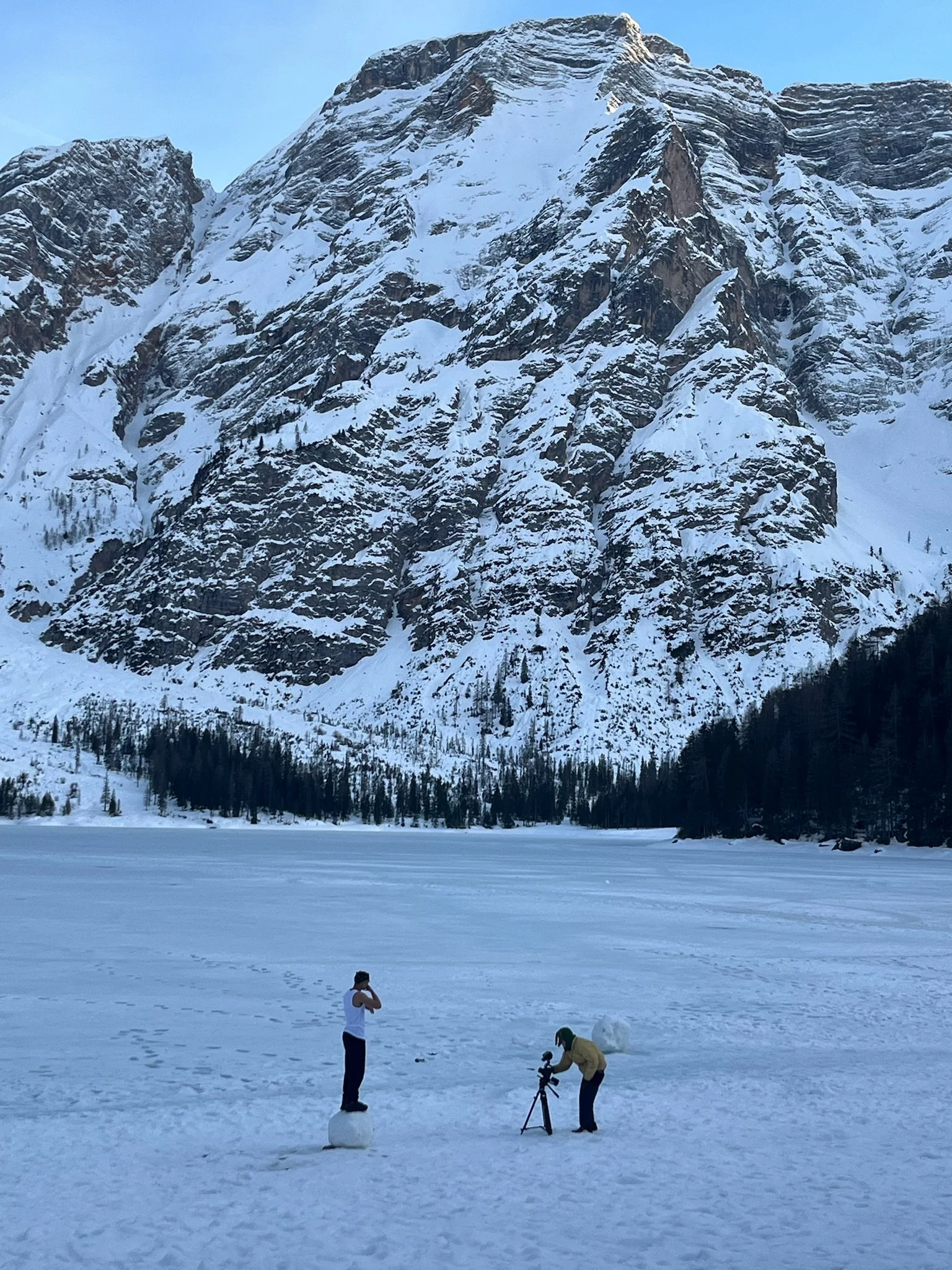 People in winter clothing on a snowy landscape with a mountain in the background; one person is standing on a small snowball, another is adjusting a camera on a tripod, and a dog is nearby.