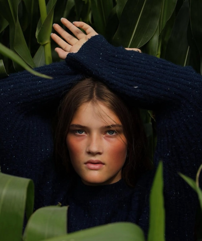 A young woman with blue eyes and brown hair, wearing a navy sweater, is surrounded by green corn plants. She has her arms crossed above her head, holding her hands together.