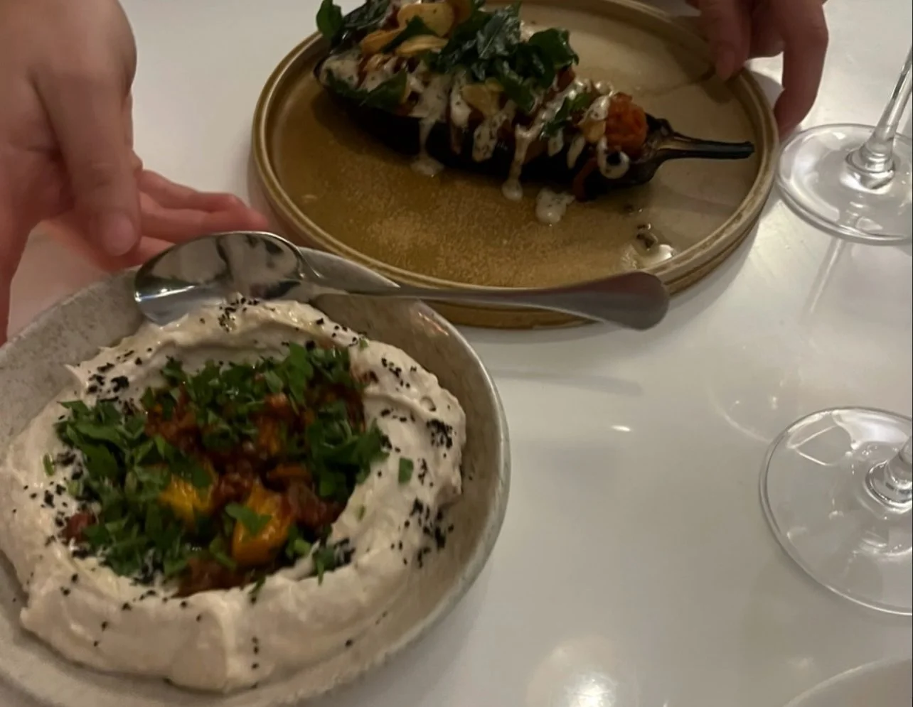 A bowl of hummus topped with chopped herbs and vegetables in front of a plate with a roasted eggplant garnished with sauce and greens, on a white table with wine glasses.