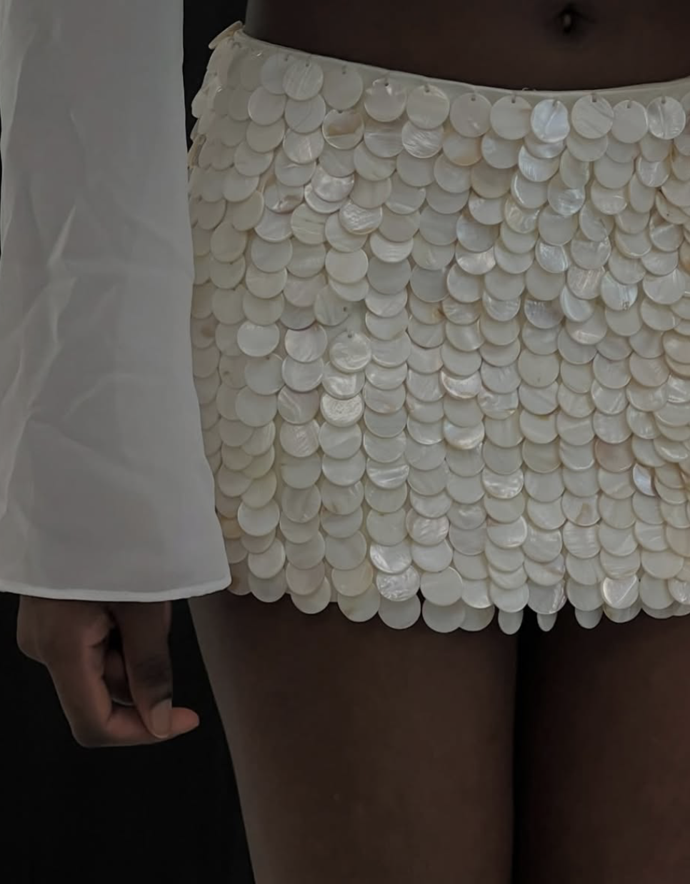 Close-up of a person wearing a cream-colored skirt made of shiny, overlapping disc-shaped sequins, with part of a white jacket visible on the left.