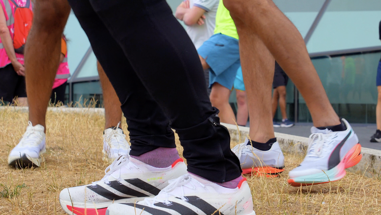 Close-up of people wearing athletic shoes, standing outdoors on dry grass, with a building and other people in the background.