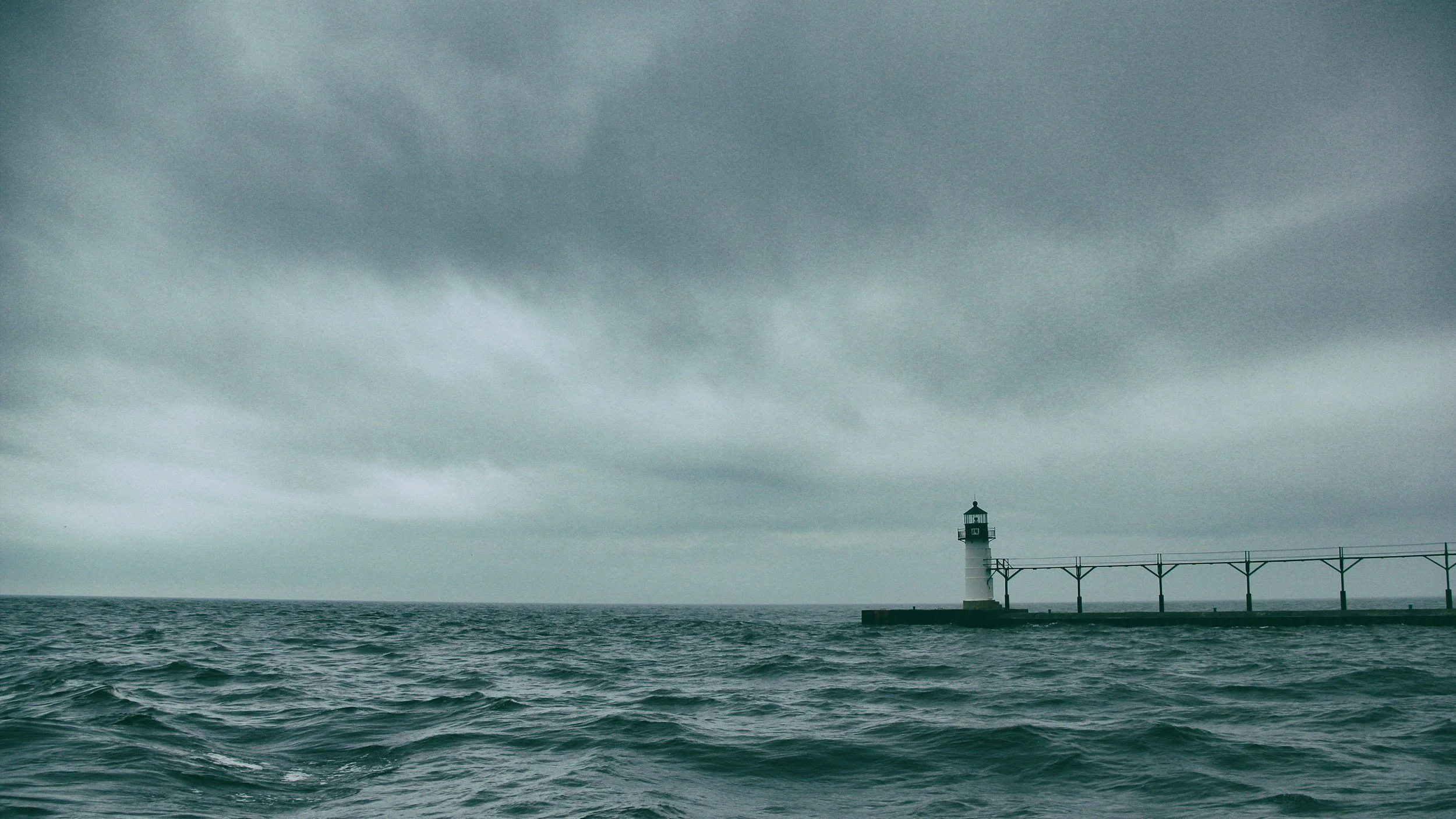 A lighthouse on a pier extending into a choppy sea under a dark, cloudy sky.