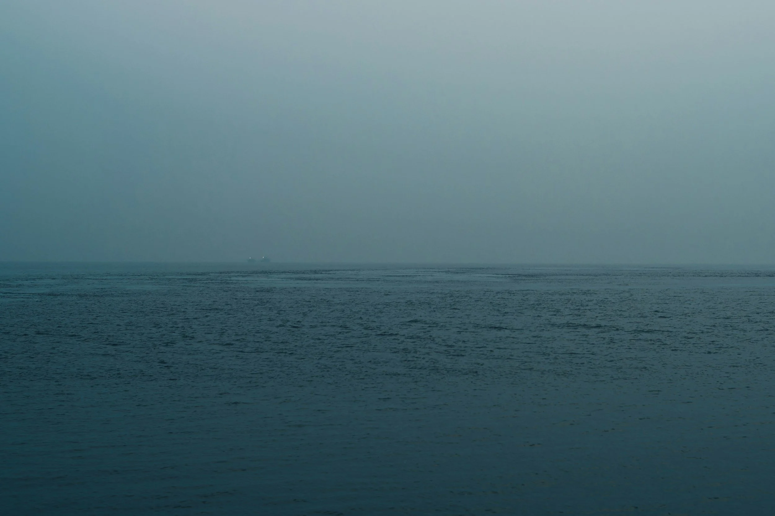 View of the ocean with calm waves and a small ship or boat in the distance under a cloudy, overcast sky.