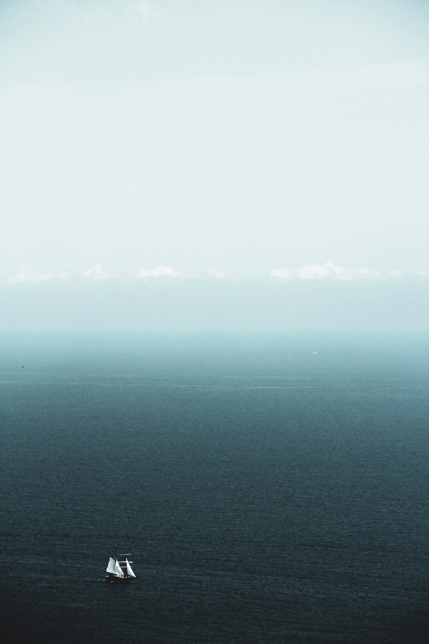 A sailboat with white sails on a dark blue ocean under a clear sky with gradient shades of blue and turquoise.