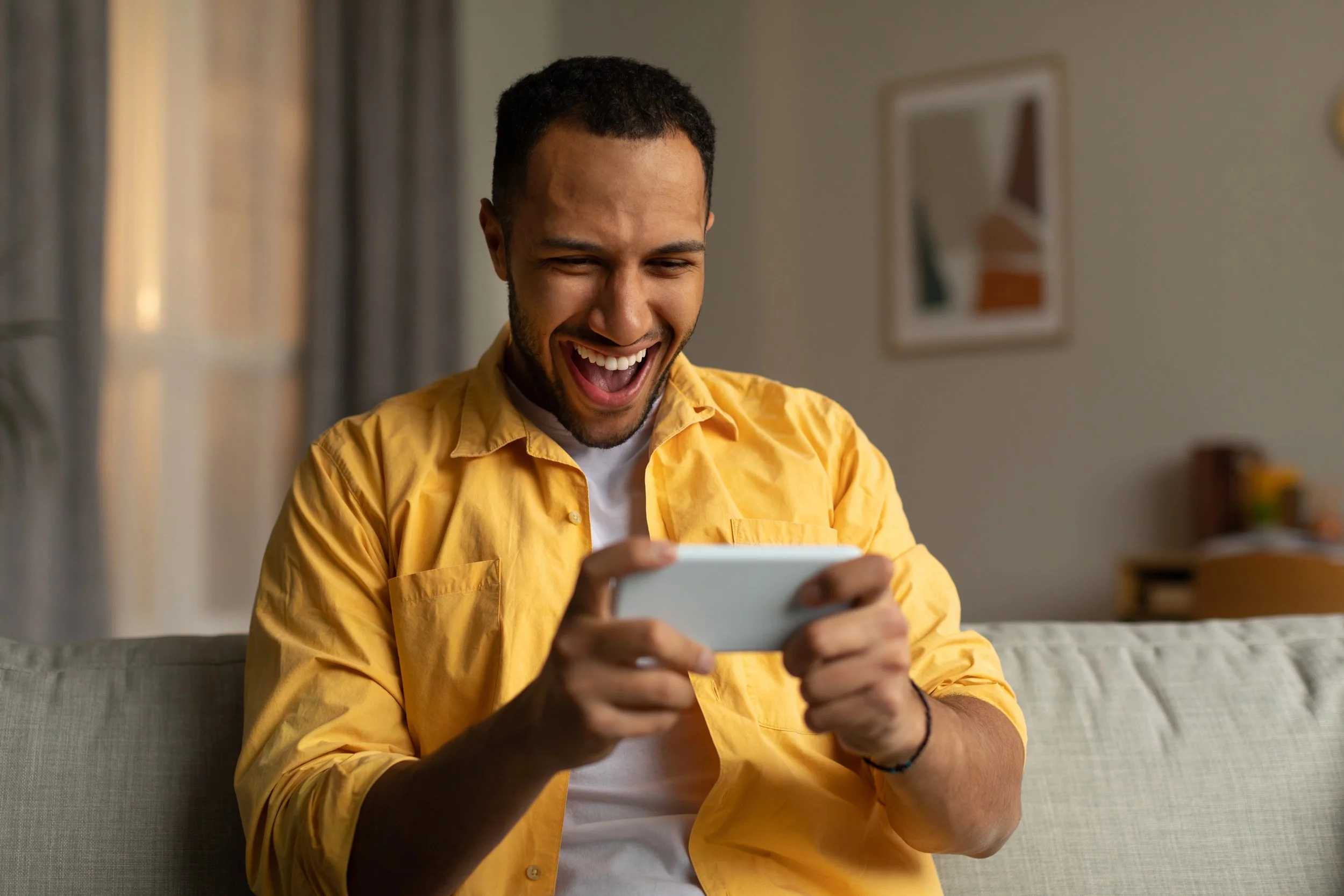 Overjoyed young black man with smartphone feeling excited over winning lottery or casino bet online, sitting on couch at home. emotional african american guy playing video game, gambling indoors