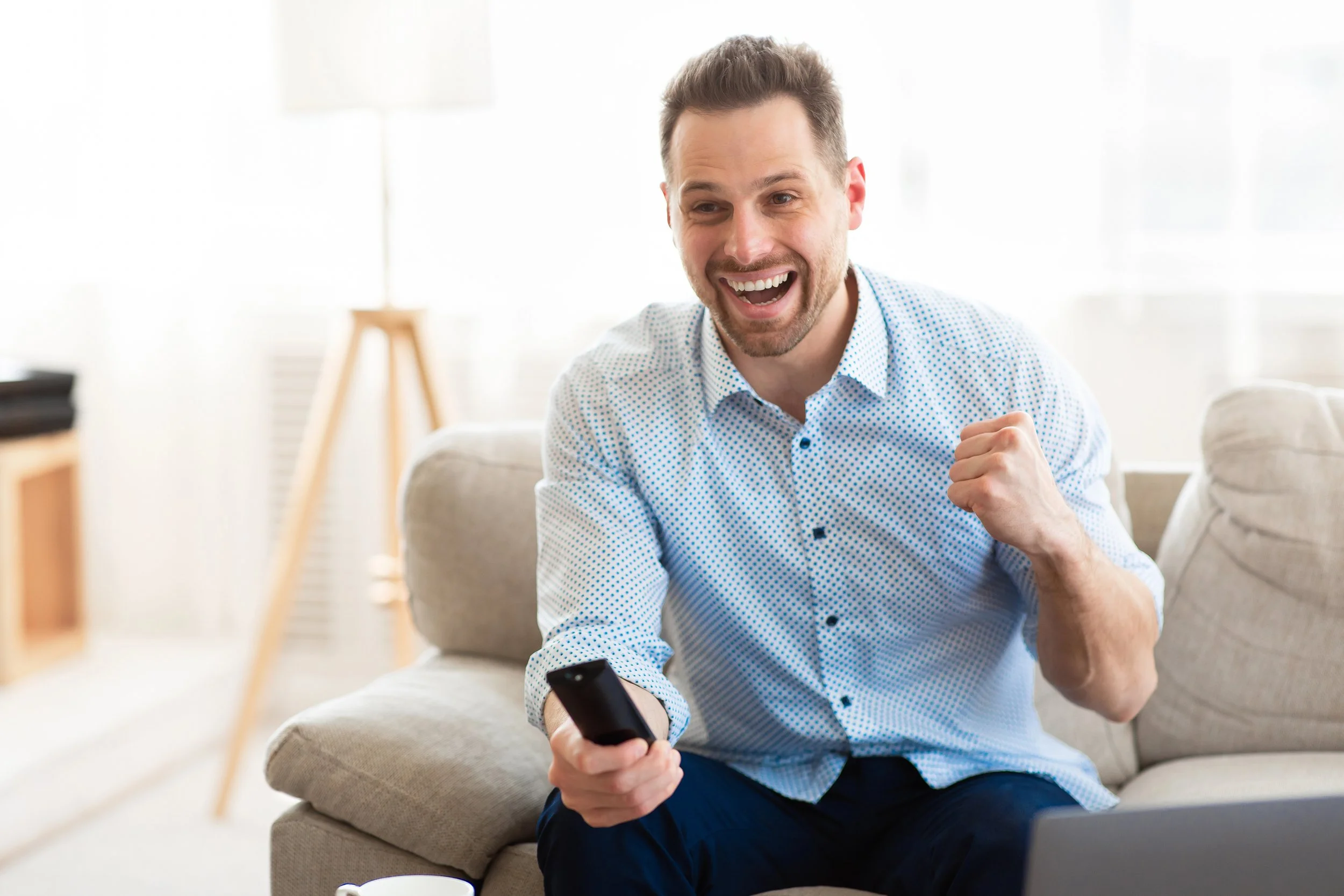 A man with short brown hair and a beard, wearing a light blue dotted shirt, sitting on a beige couch, laughing while holding a remote control in his hand.