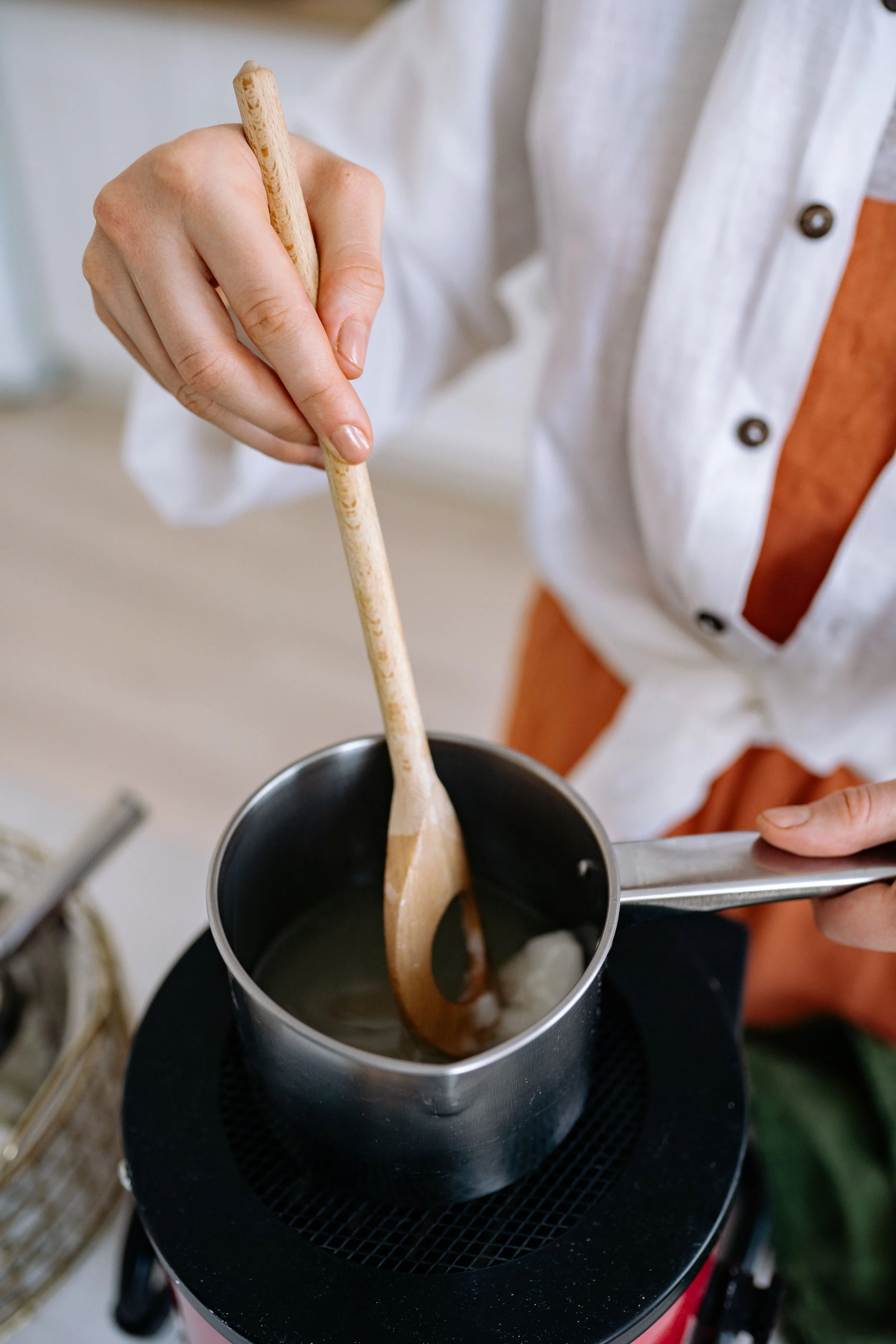 Person stirring melted wax using a wooden spoon, with a portable burner underneath, in a kitchen setting.