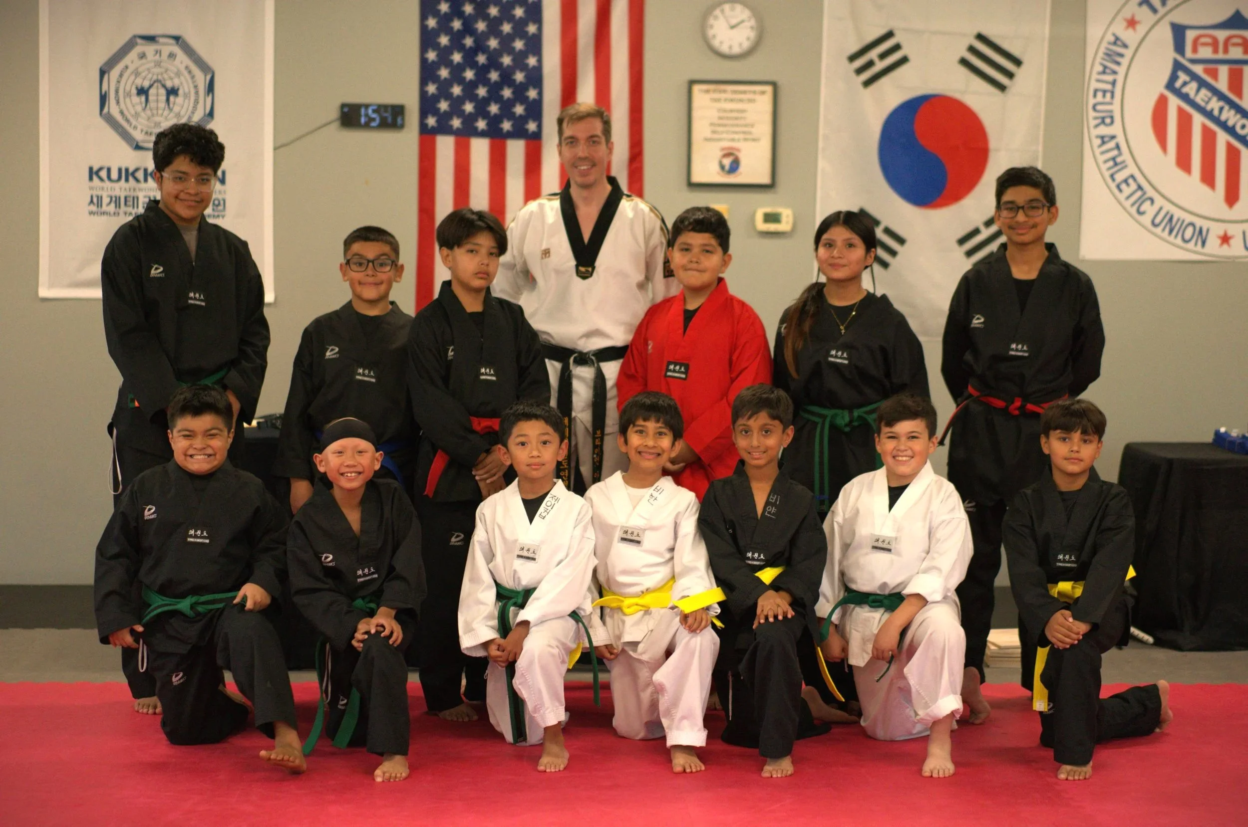 A group of children and an instructor in martial arts uniforms posed for a group photo in a martial arts dojo. The background features flags of the United States, South Korea, and Korean Taekwondo flags, along with banners and a clock.