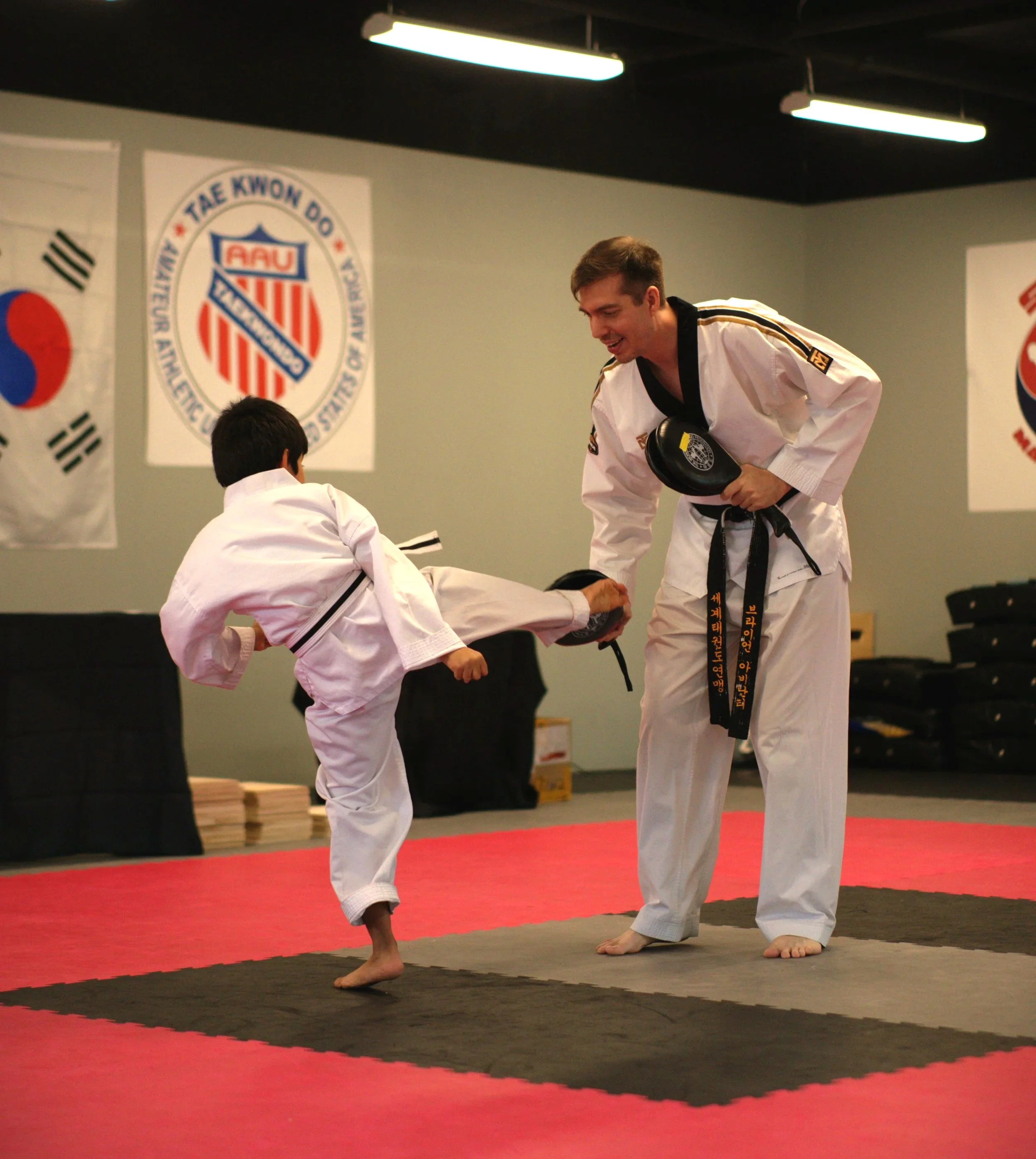 Two martial artists in gis practicing Taekwondo in a dojo, with US and South Korean flags and a Taekwondo USA emblem on the wall.