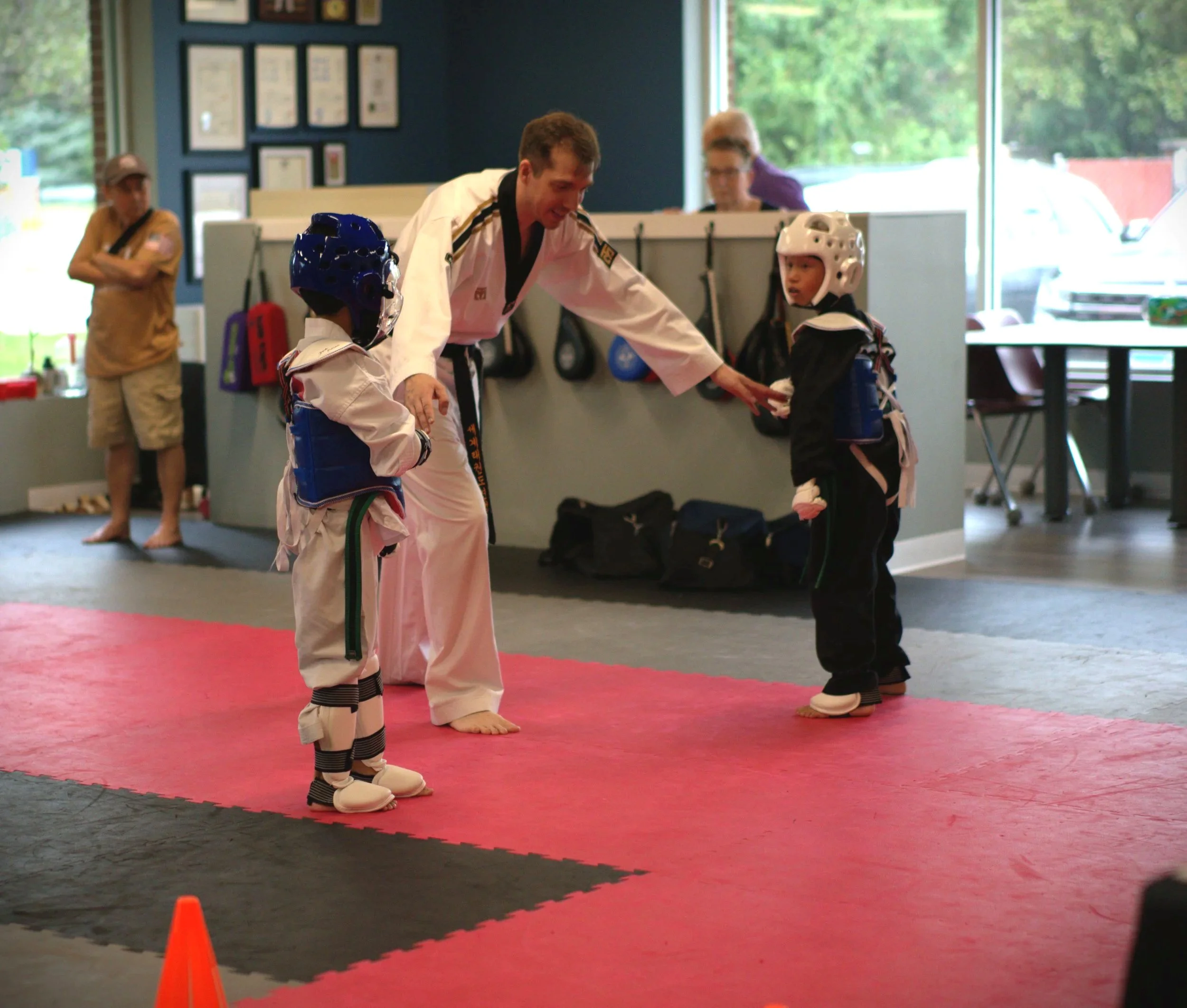 A martial arts instructor teaching two young children wearing protective gear in a dojo with large windows and framed certificates on the wall.