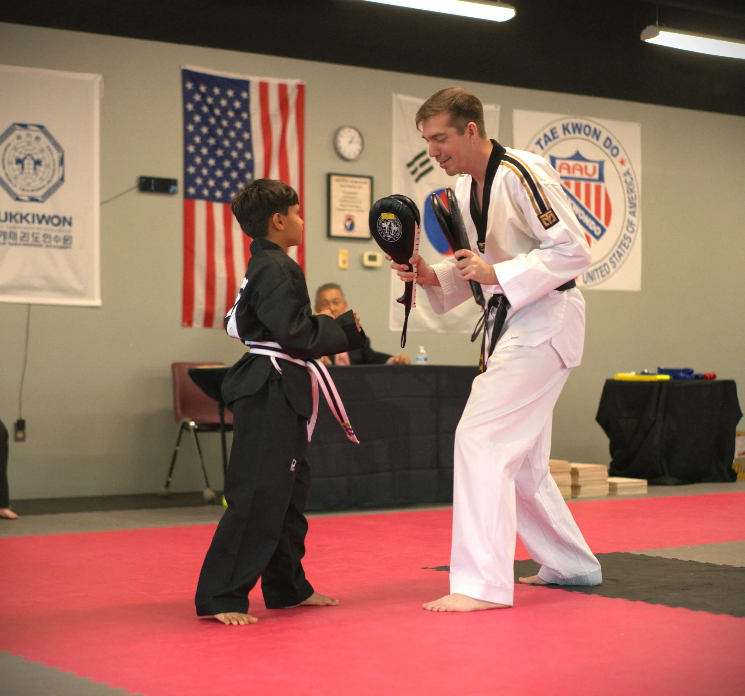 A martial arts instructor and a young student in a dojo practicing a technique. The instructor is wearing a white gi and black belt, holding focus mitts, while the student is dressed in black gi with a pink belt.