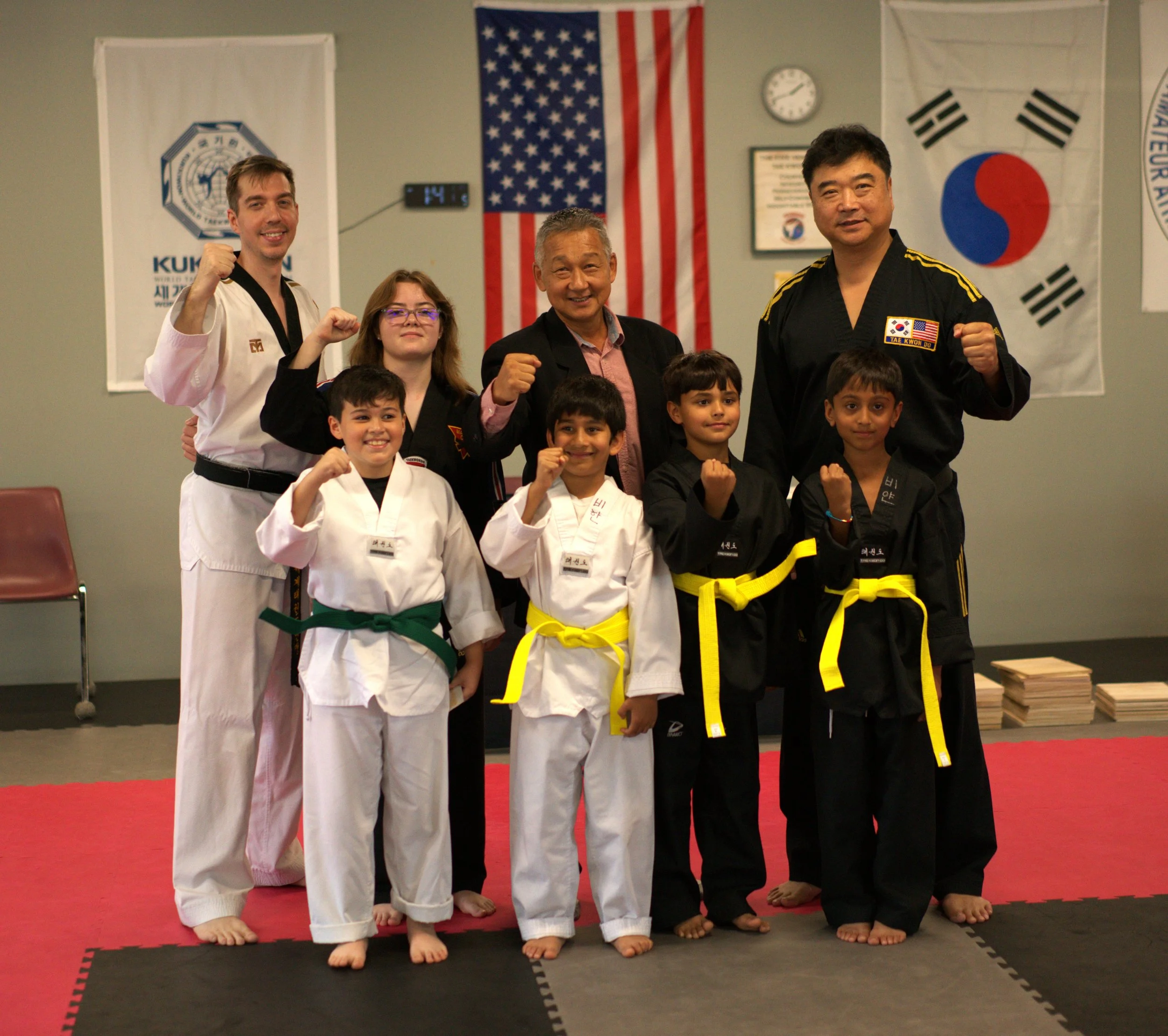 A group of martial artists and a instructor posing together in a dojo, some in white gis with colored belts, others in black gis with yellow belts, standing on red and black mats. They are making fist gestures. The background displays flags of the Un