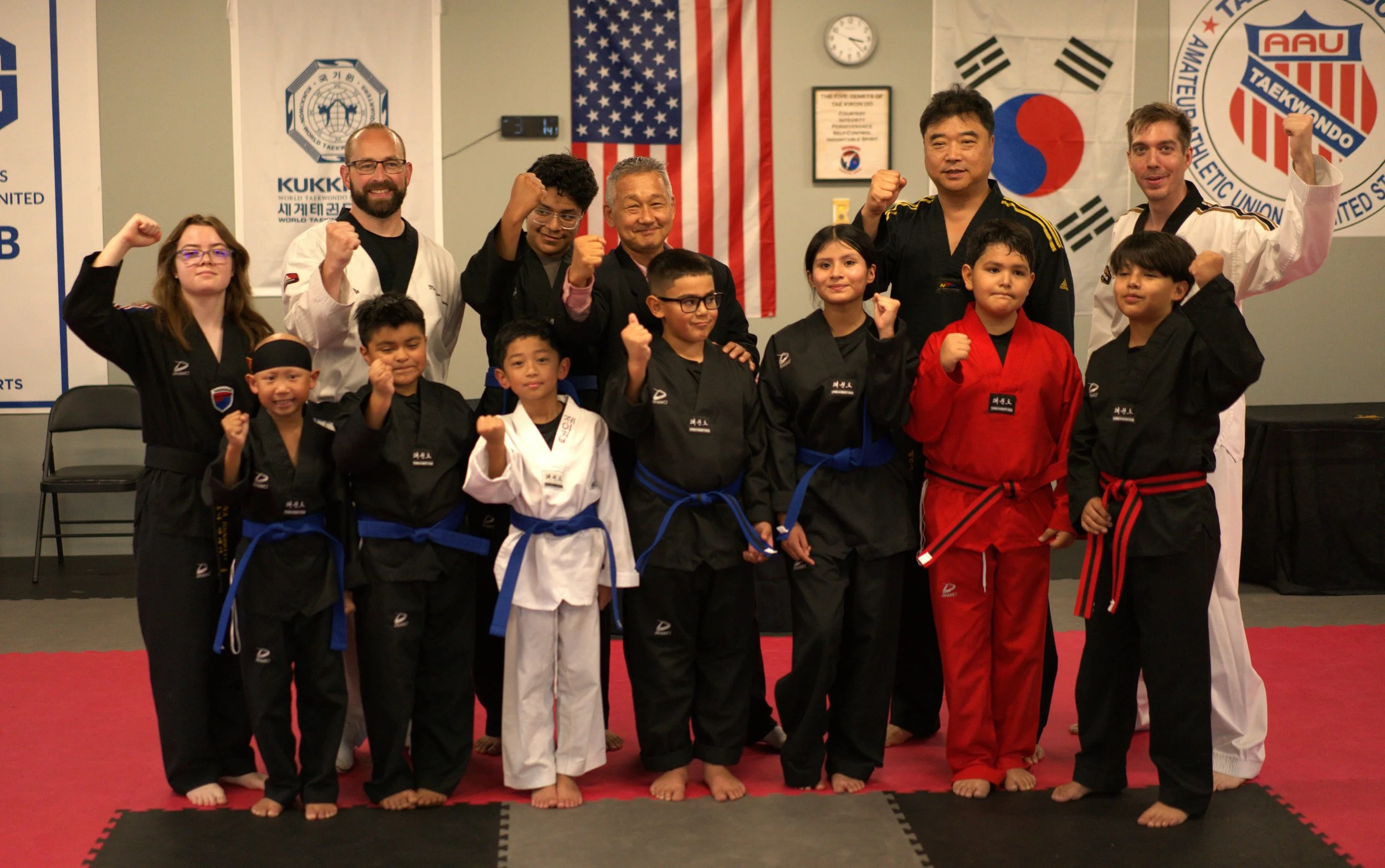 A group of martial artists and instructors in uniform, some in black, red, and white belts, standing on a martial arts mat with fists raised. The background features flags, including the American flag, the South Korean flag, and banners with martial 