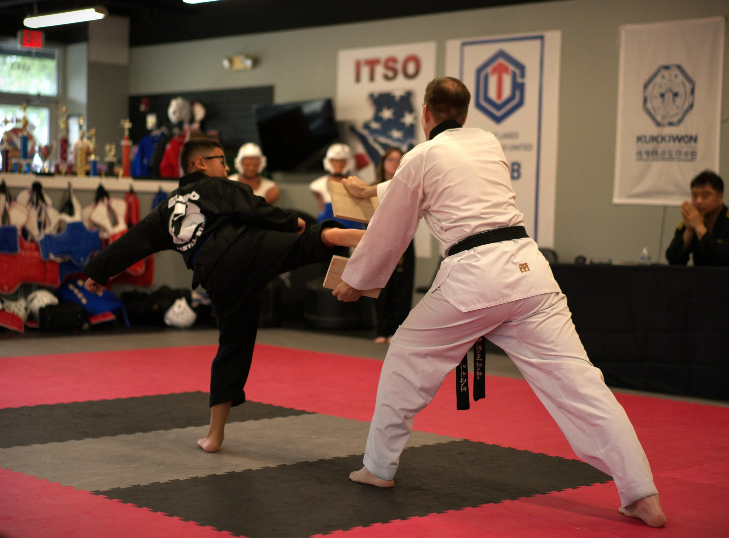 A martial arts instructor in a white gi with a black belt demonstrating a technique to a young student in a black gi with a black belt in a martial arts dojo. The dojo has banners and trophies on display.