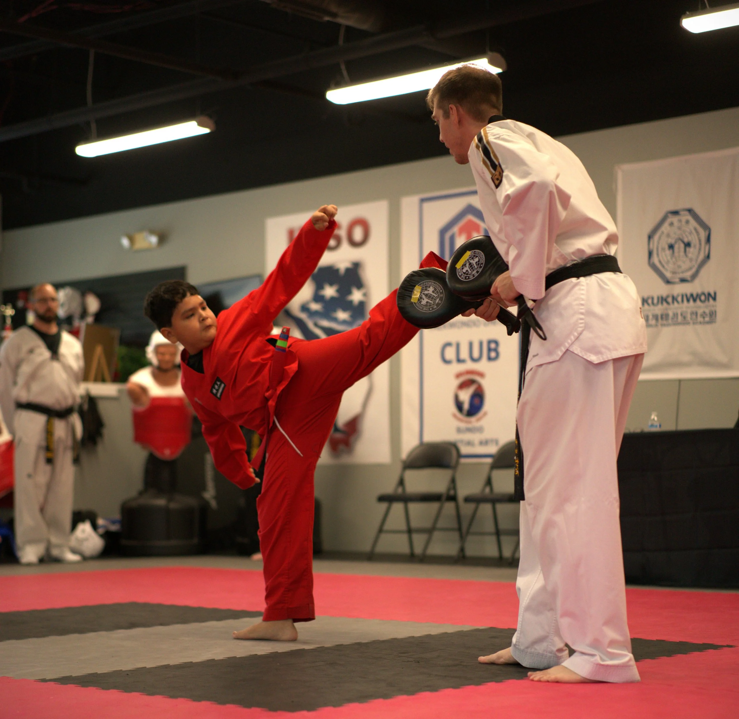 Two young martial artists bowing to each other in a martial arts training dojo, one wearing a red gi and the other wearing a white gi, with a man in the background observing.