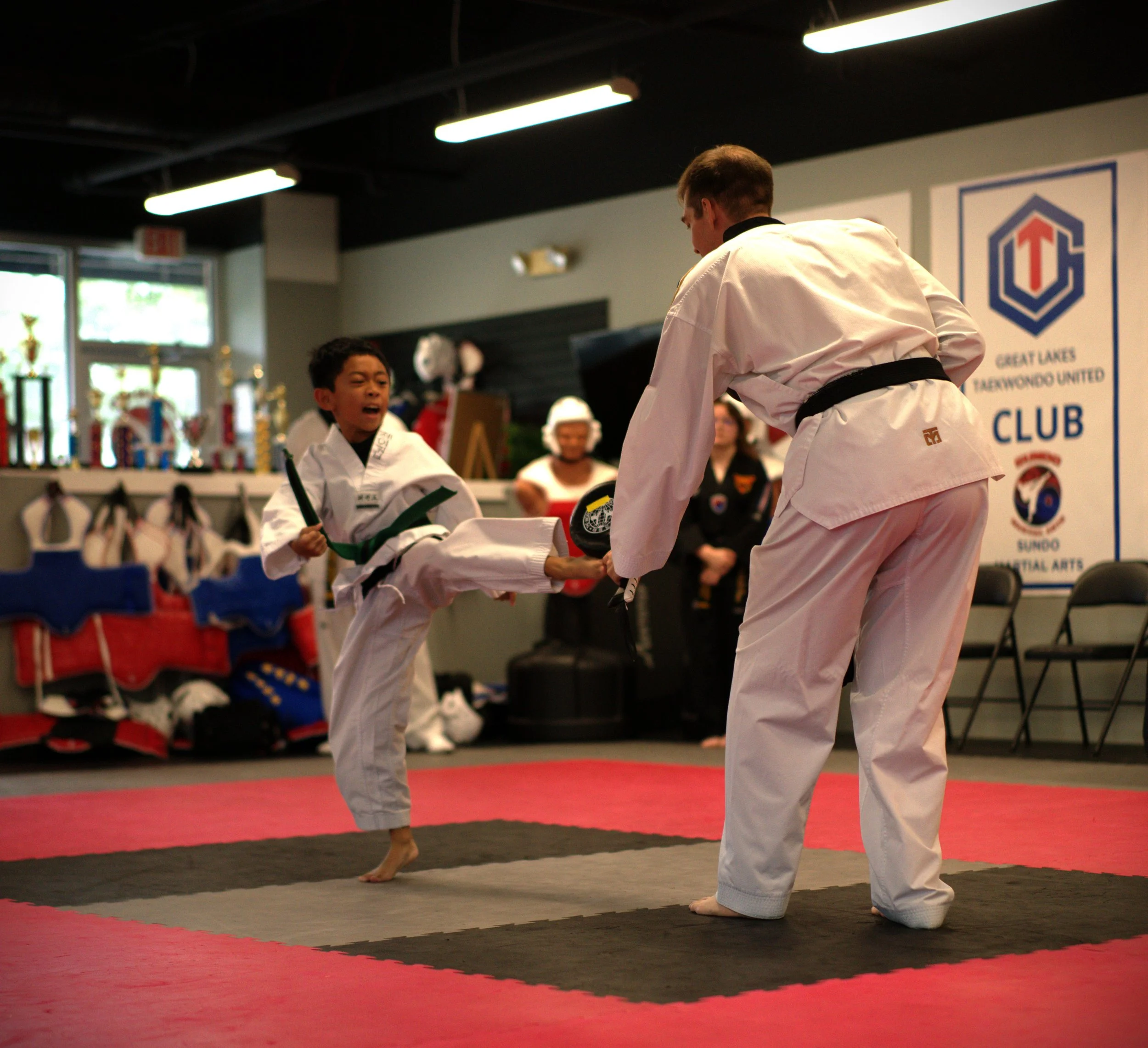 Two young martial artists practicing taekwondo in a dojo, wearing traditional white uniforms and black belts, with trophies and a banner in the background.