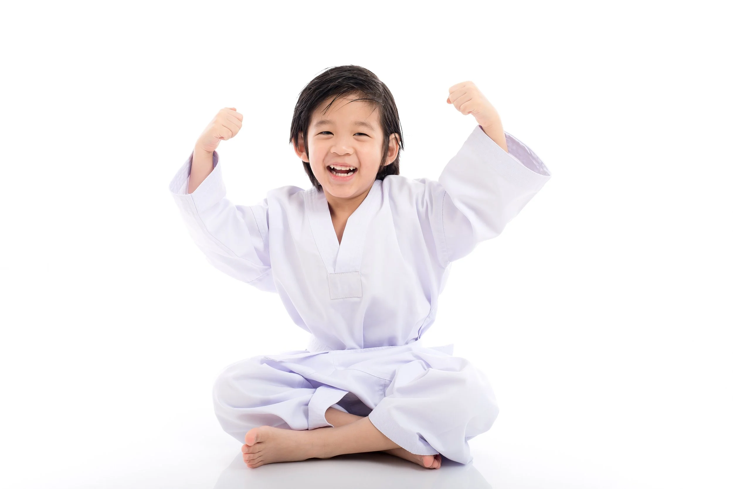 A young child in a white martial arts uniform sitting cross-legged and smiling, with arms raised and fists clenched in a victory pose.