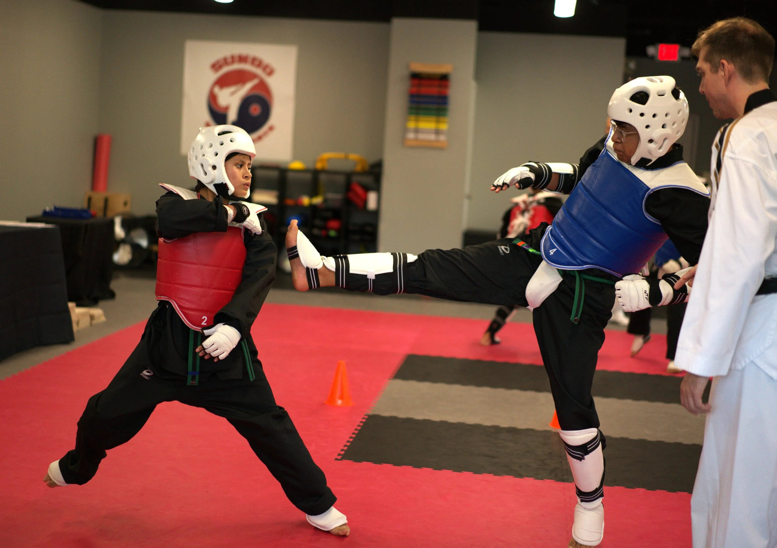 Two young martial artists are practicing a sparring session with the instructor at a martial arts dojo. One is wearing red protective gear and is in a fighting stance, while the other is wearing blue protective gear and is executing a kick.