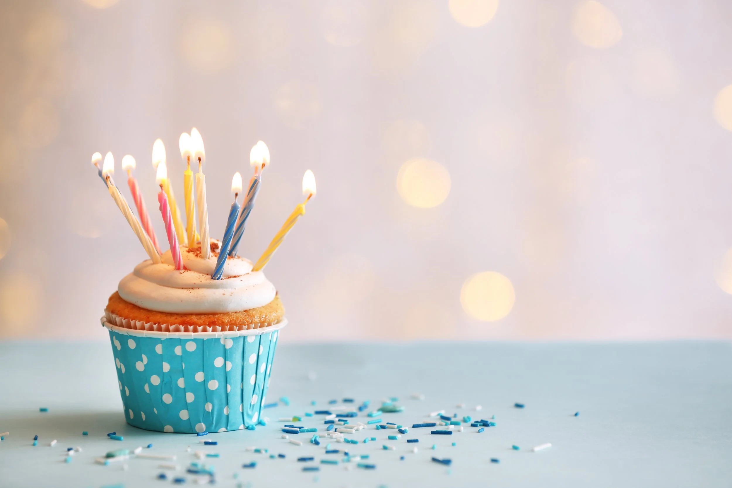 Cupcake with white frosting and colorful birthday candles in a blue polka dot wrapper, with scattered blue and white confetti on a light blue surface, and bokeh lights in the background.