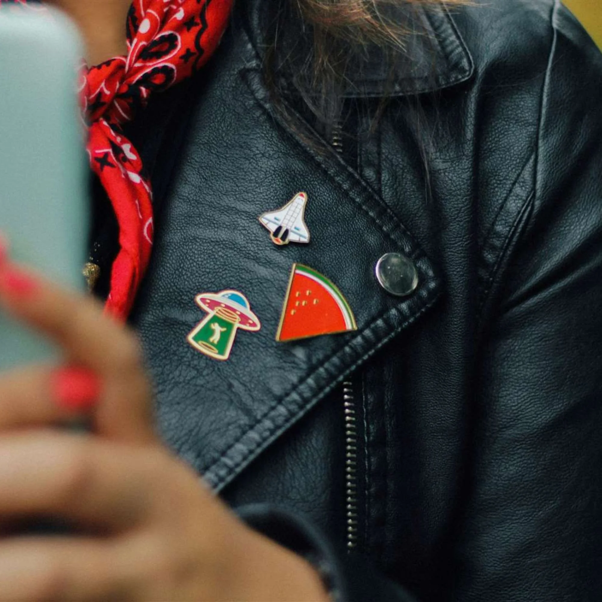 Enamel pin badges from The Pin Joint, featuring a space shuttle, UFO, and watermelon slice, displayed on a black leather jacket.