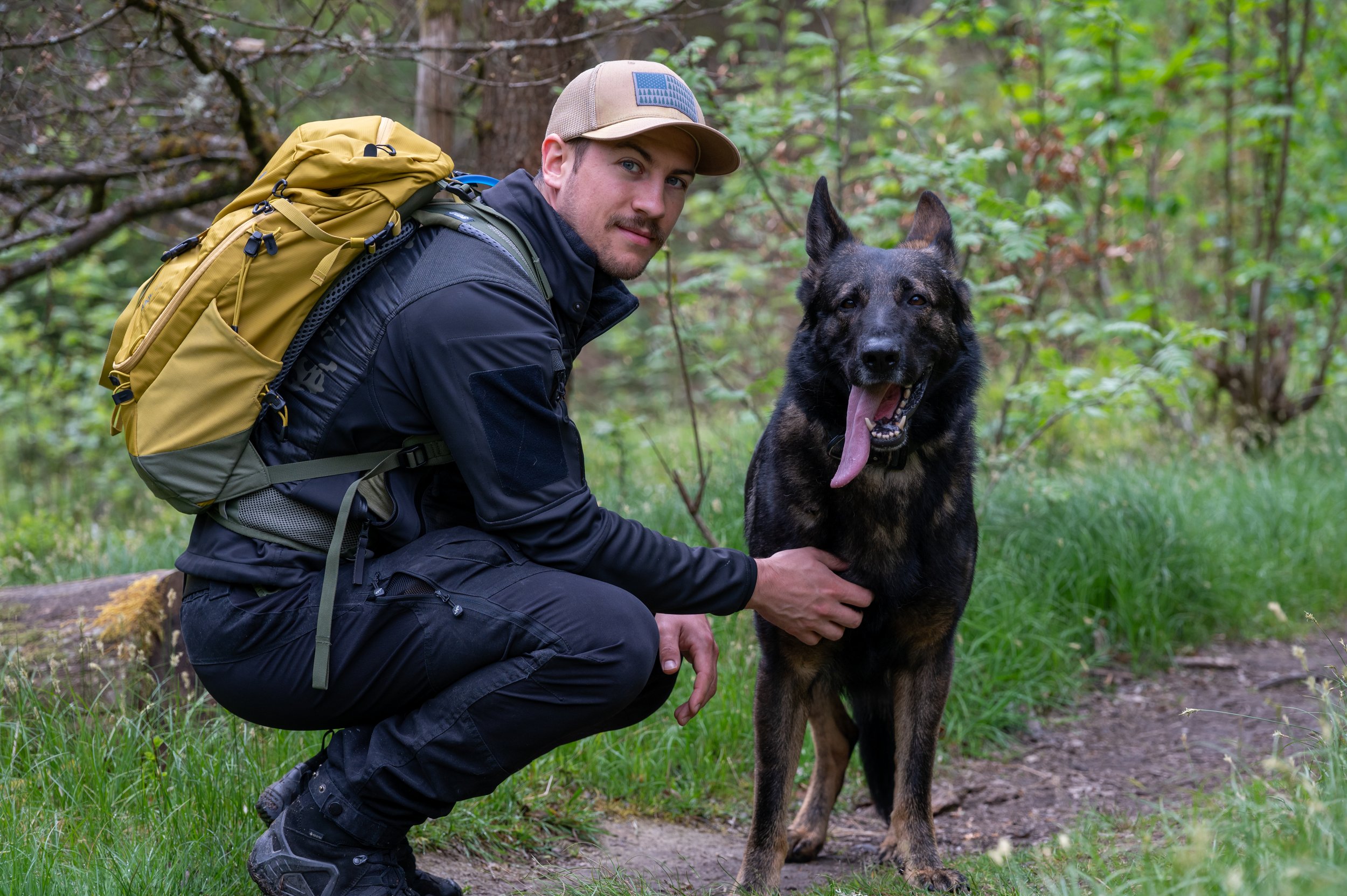 Ein Mann in dunkler Outdoor-Kleidung mit gelbem Rucksack und Cap kniet neben einem deutschen Schäferhund in einem grünen Wald.