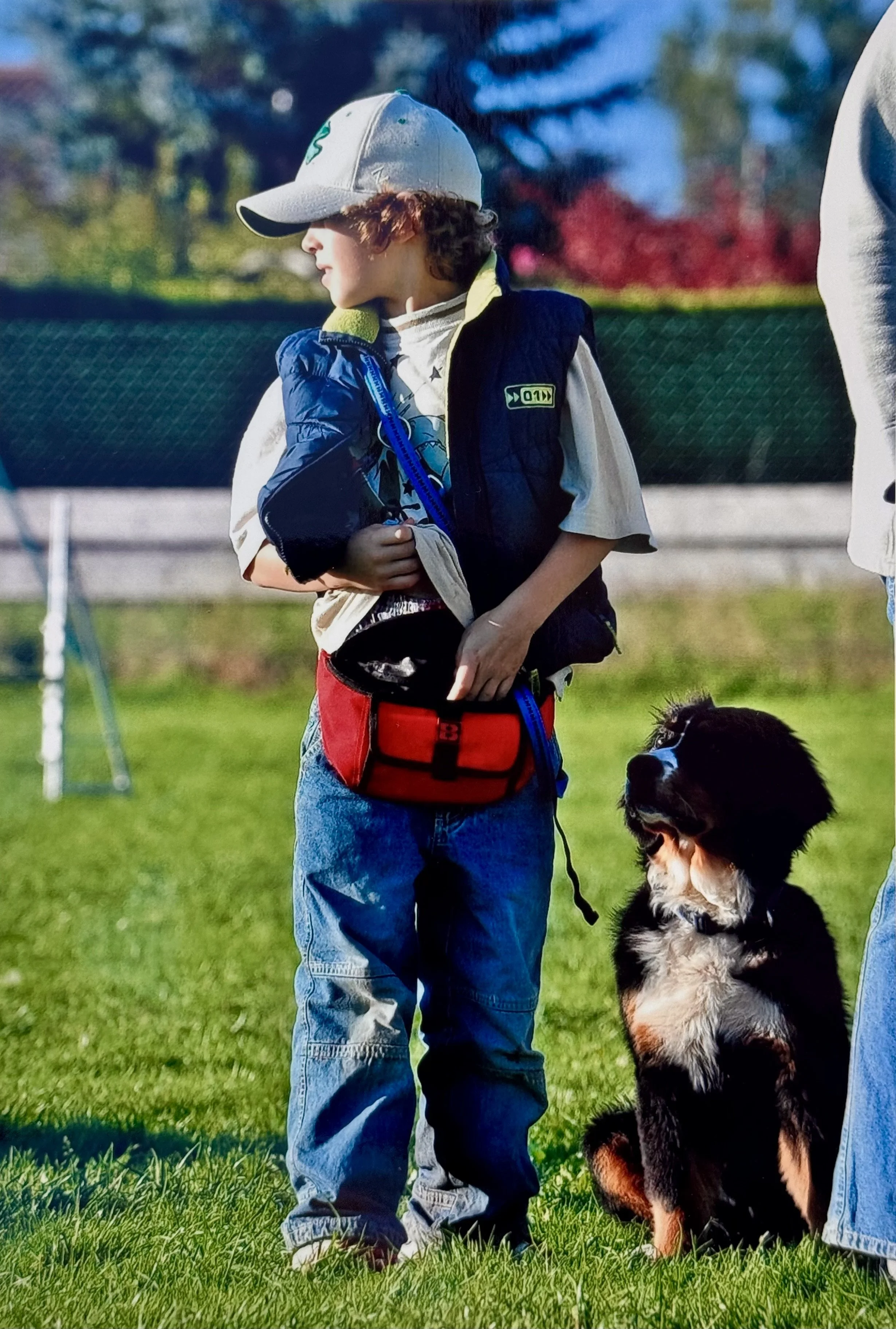 Ein Junge mit braunen lockigen Haaren trägt eine graue Cap und eine schwarze Weste, steht auf grünem Rasen, mit einem Bernhardiner-Hund zu seiner Rechten.