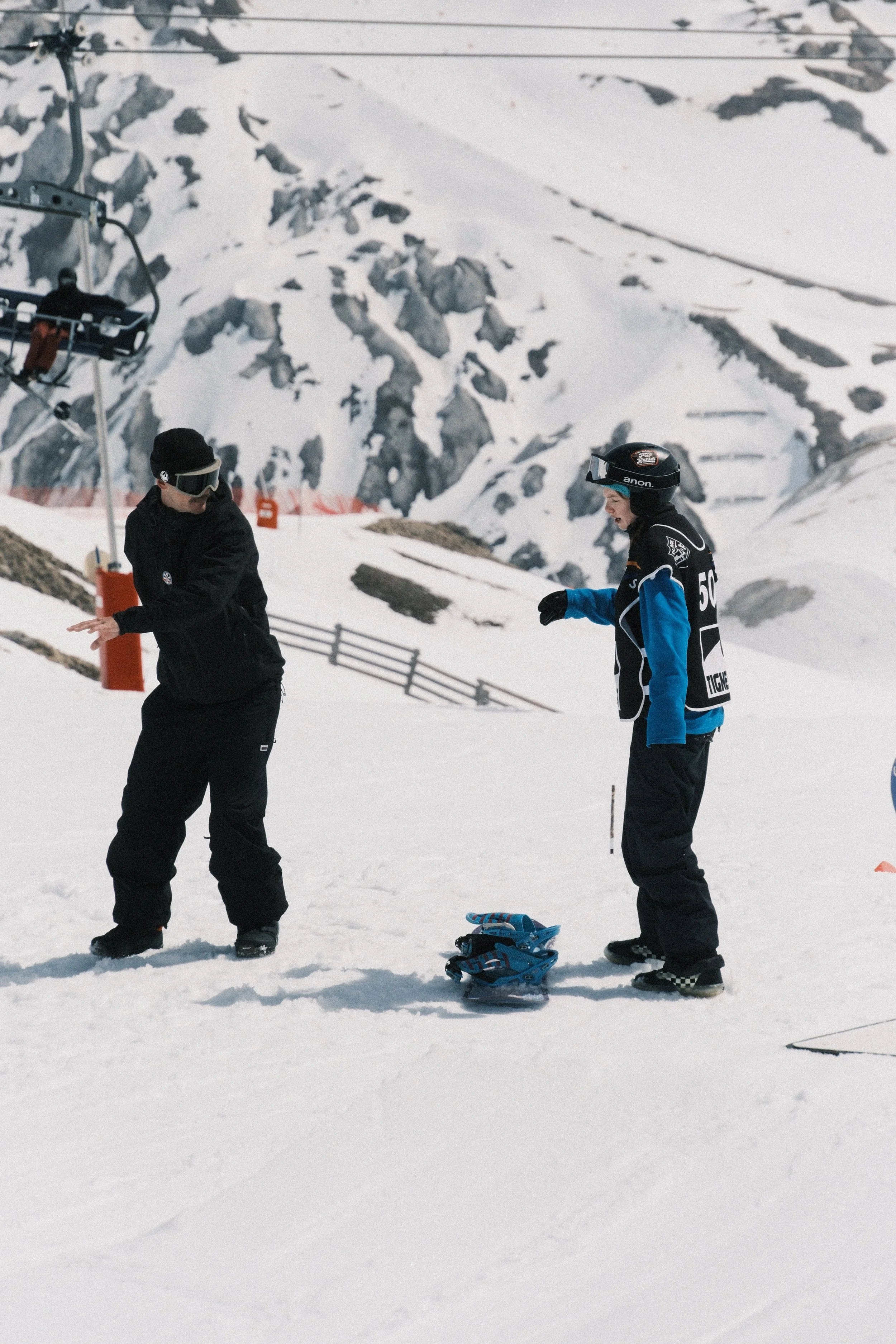 Two snowboarders stand on snow in a mountain ski resort, with one adjusting their equipment and the other looking down, while a ski lift and rocky, snow-covered mountain slopes are visible in the background.