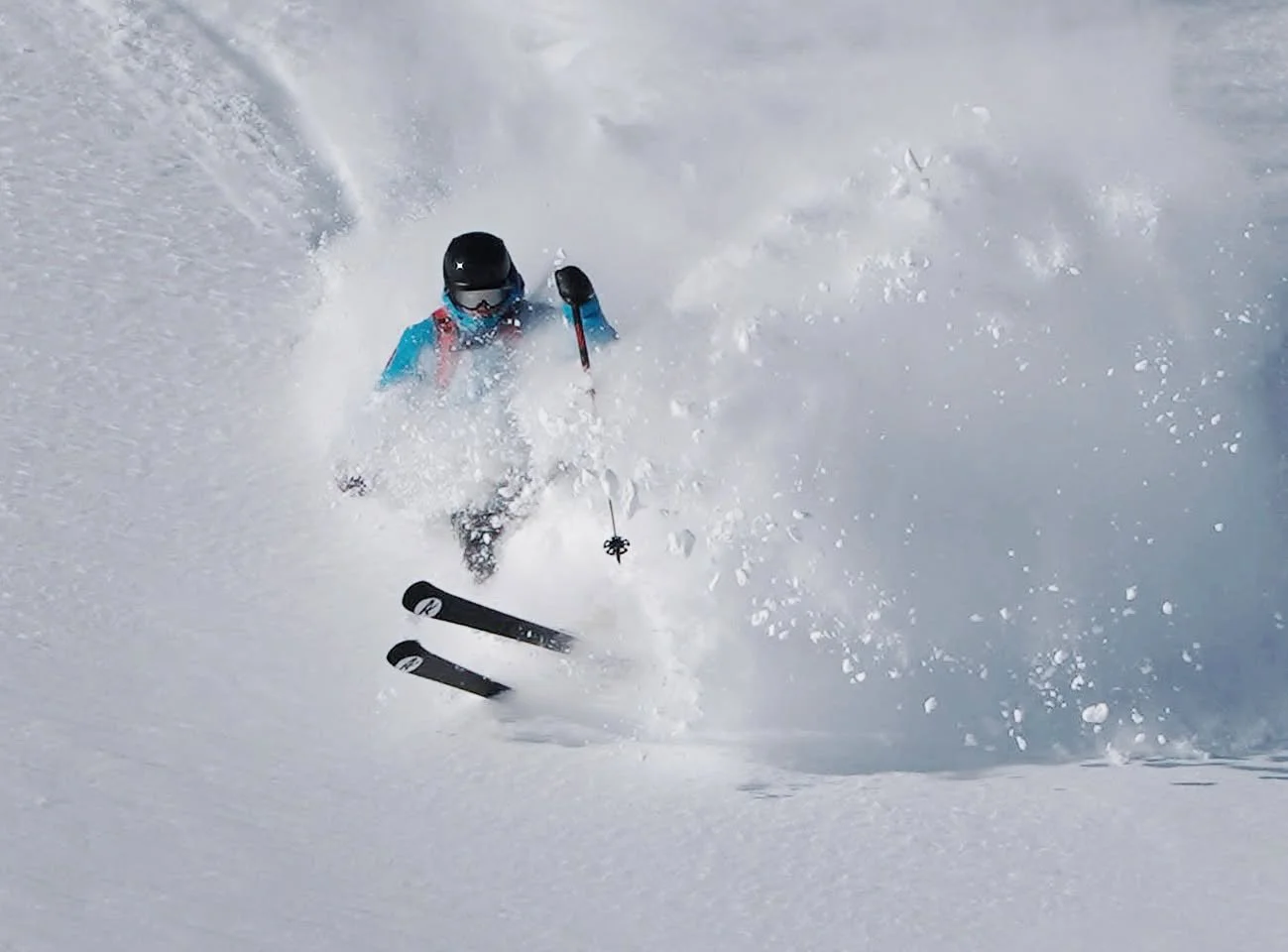 Person skiing in deep snow, wearing a black helmet, goggles, a blue jacket, and black skis, creating a snow spray.