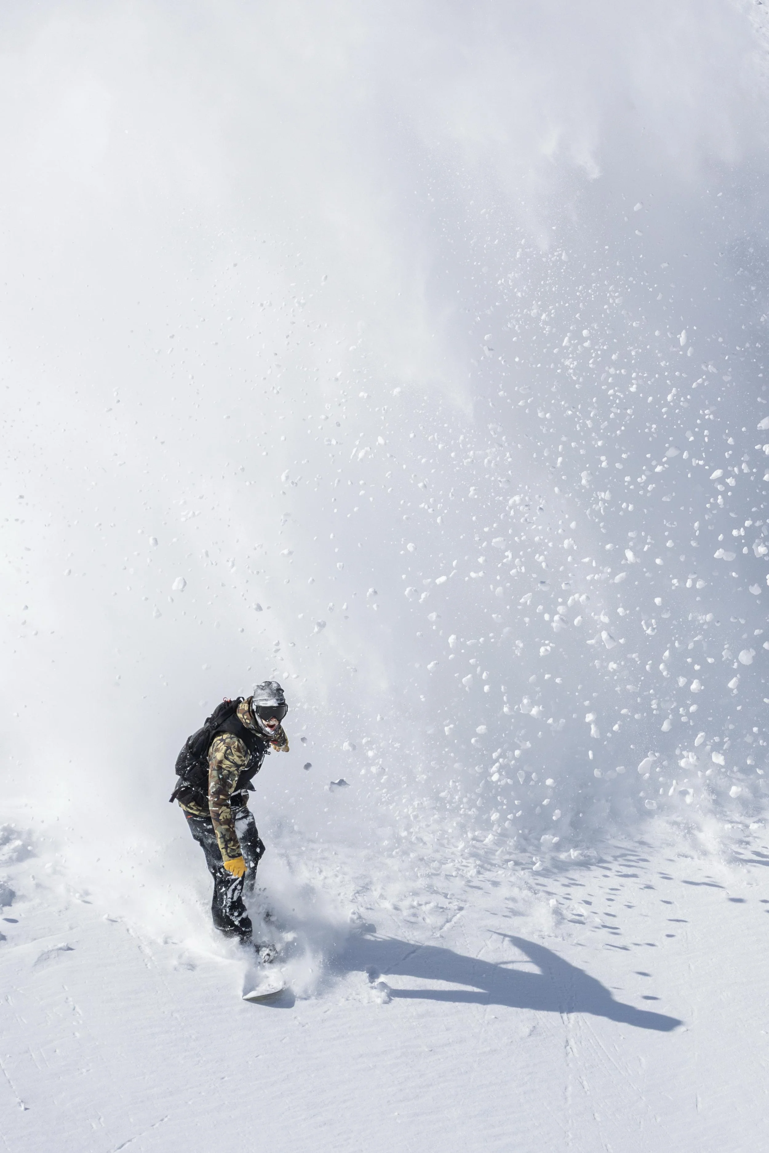 Person snowboarding in snowy mountain landscape with a cloudy sky, wearing camouflage clothing, gloves, and goggles.