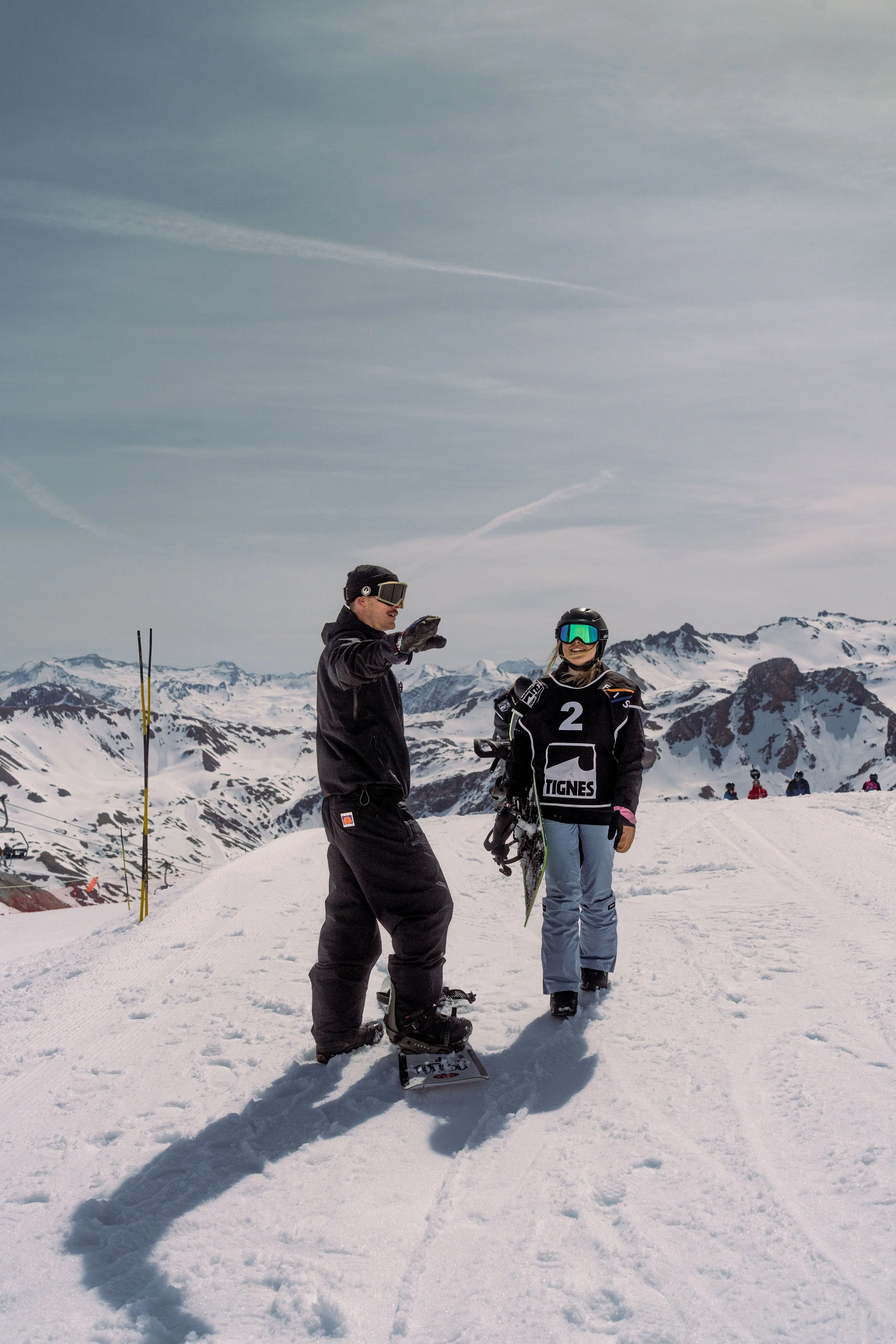 Two people standing on a snowy mountain landscape, one wearing snowboard gear and the other with a helmet and goggles, with a backdrop of snow-covered mountains and a cloudy sky.