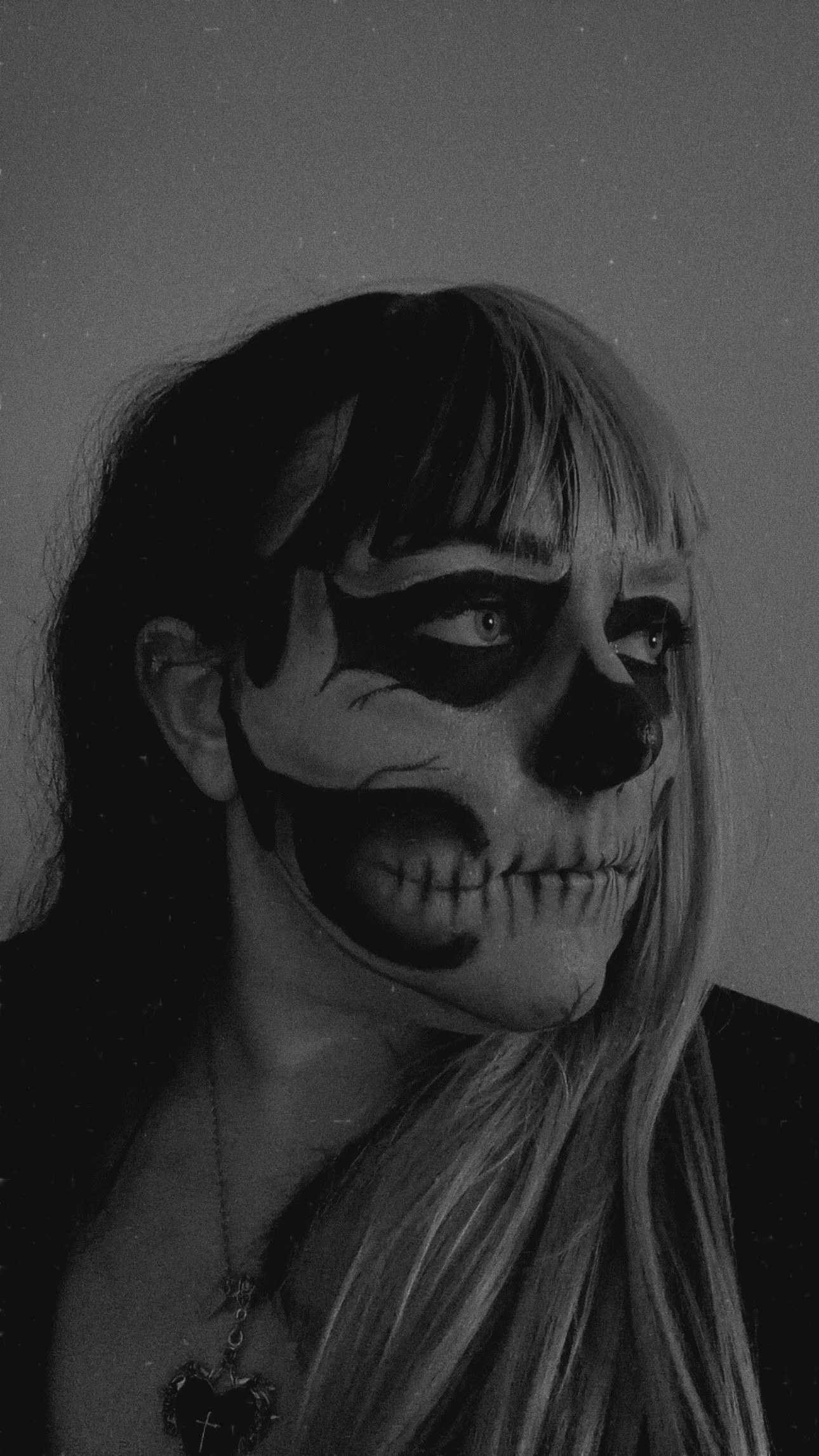Black and white photo of a woman with face makeup resembling a skull, looking to her left with long hair and wearing a necklace with a cross pendant.