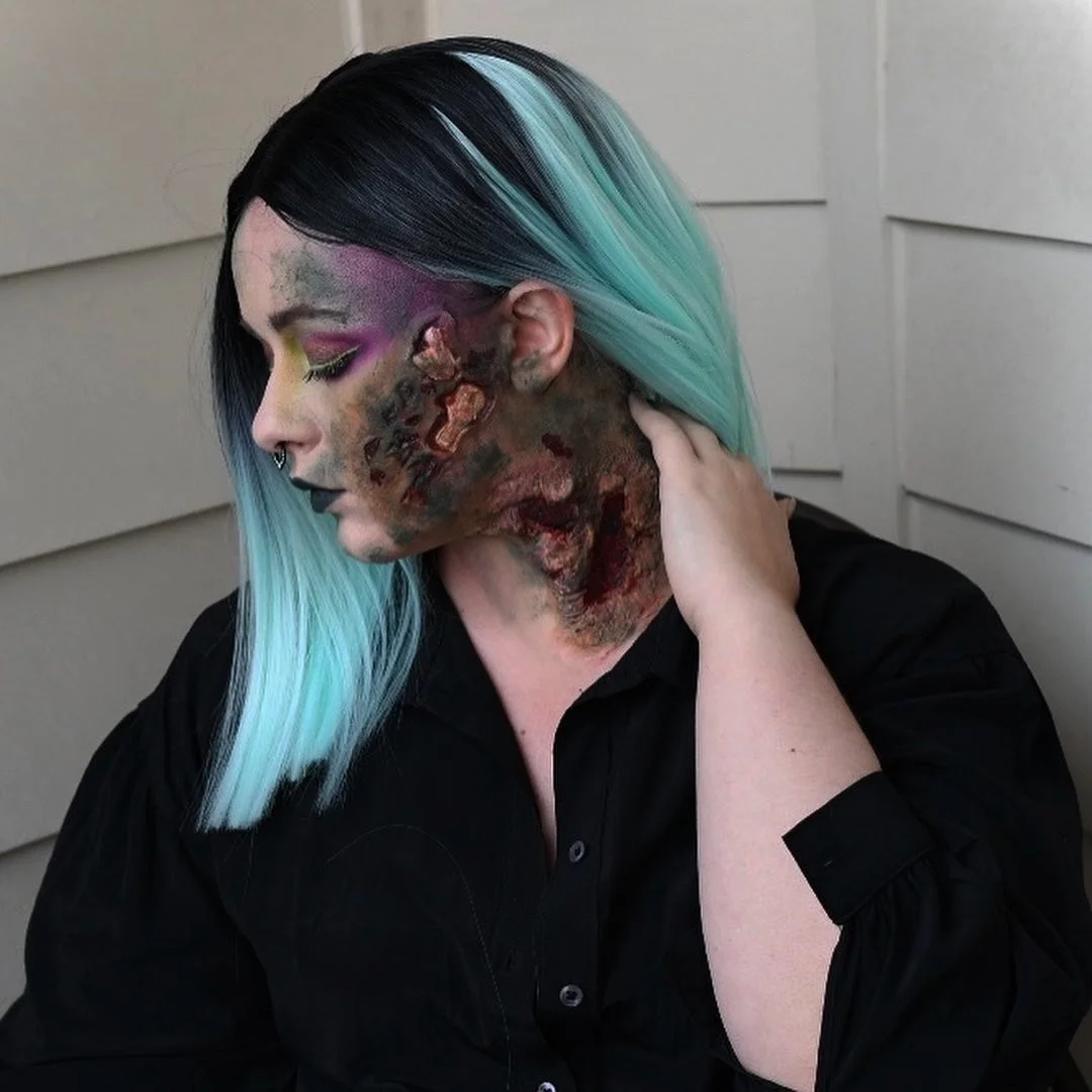 Person with black and blue hair, makeup with vibrant colors, and special effects makeup resembling burns or decayed skin on their face and neck.