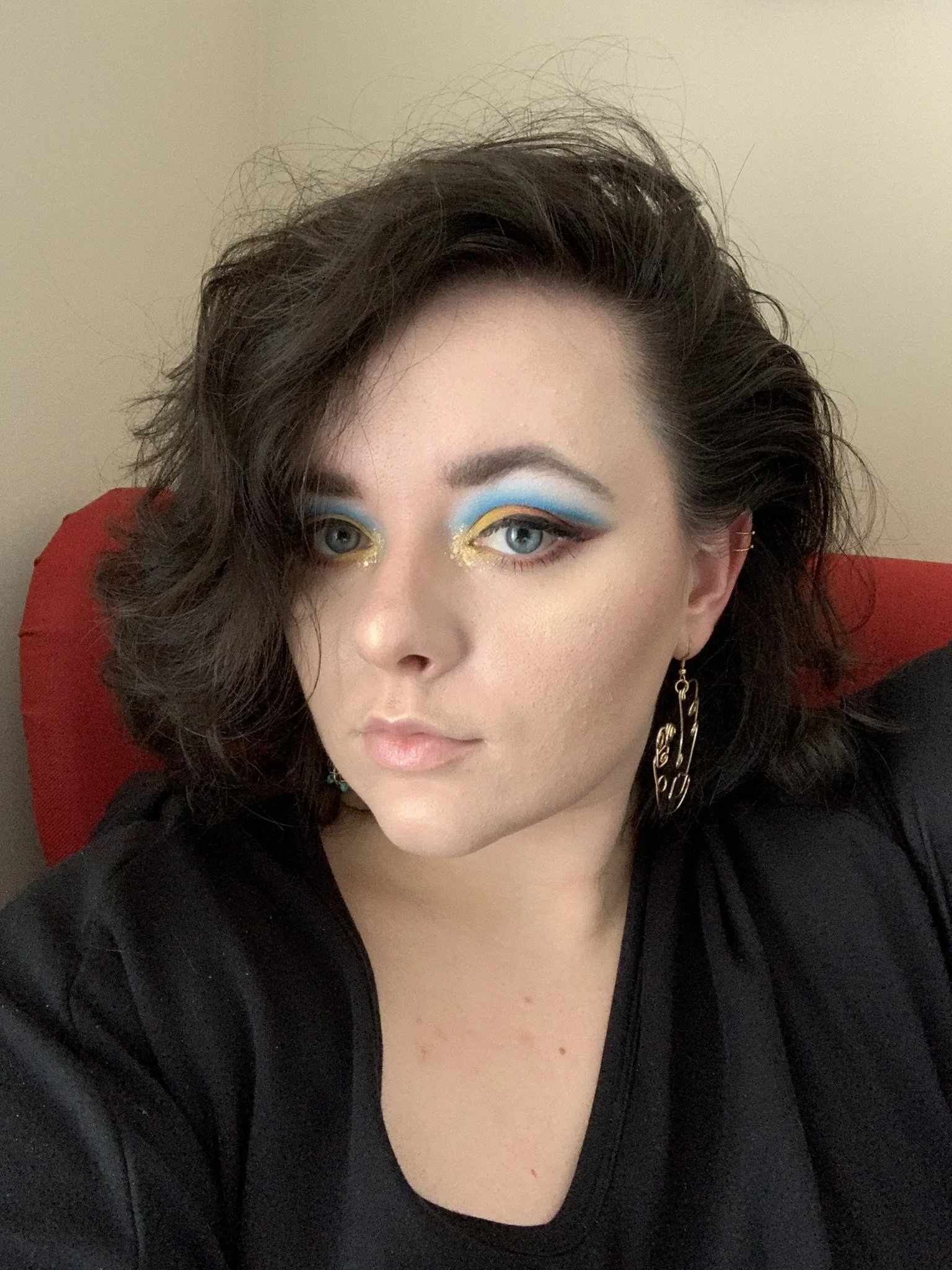 A woman with dark, curly hair and blue eyes has colorful, glittery eye makeup with yellow, blue, and black eyeshadow. She is wearing large gold earrings and a black top, sitting on a red chair against a beige background.