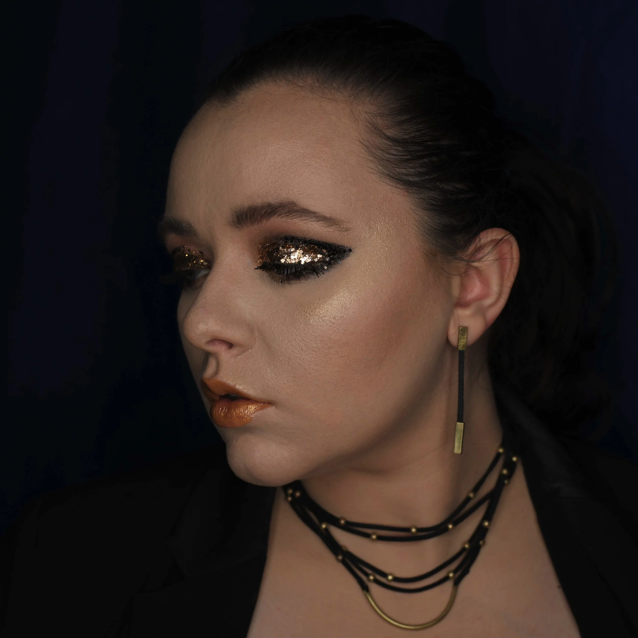 Close-up of a woman's face with dark hair, glittery eye makeup, and layered necklaces, with a dark background.