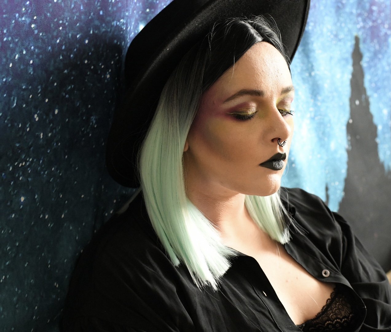 A woman with black and mint green ombre hair, wearing dark makeup including black lipstick, and a black hat, leaning against a speckled blue wall with a misty cityscape silhouette in the background.