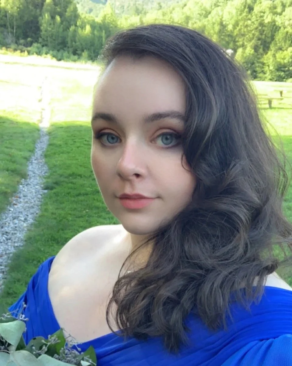 A young woman with blue eyes and long, dark, curly hair stands outside on a sunny day, with a grassy field and trees in the background, wearing an off-the-shoulder blue dress.