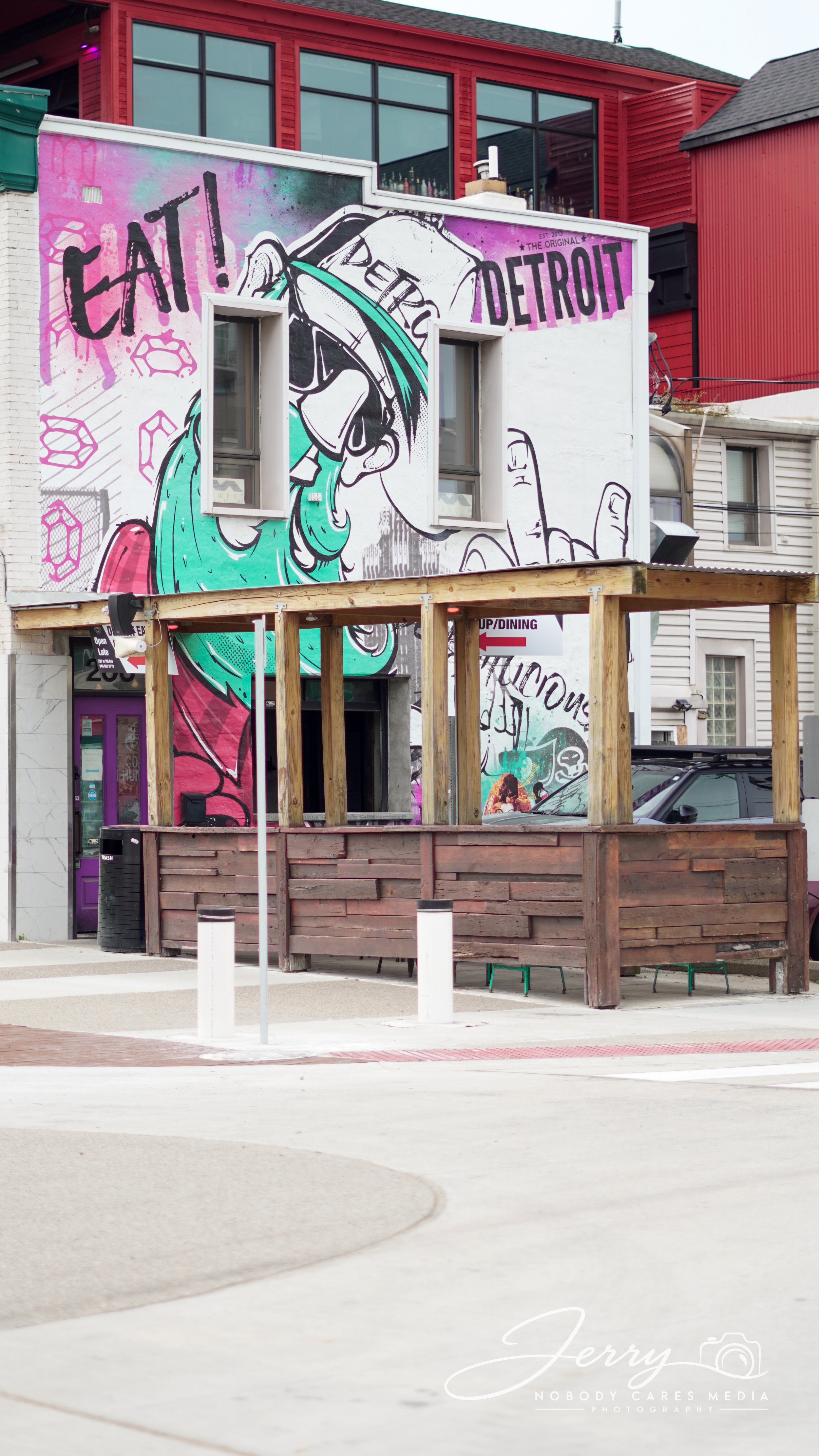 Street scene featuring a large colorful mural on the upper wall of a building, depicting a cartoon character with sunglasses and flowing hair, with the words 'EAT DETROIT' on the mural. There is a wooden deck structure in the foreground, and neighboring buildings are visible.