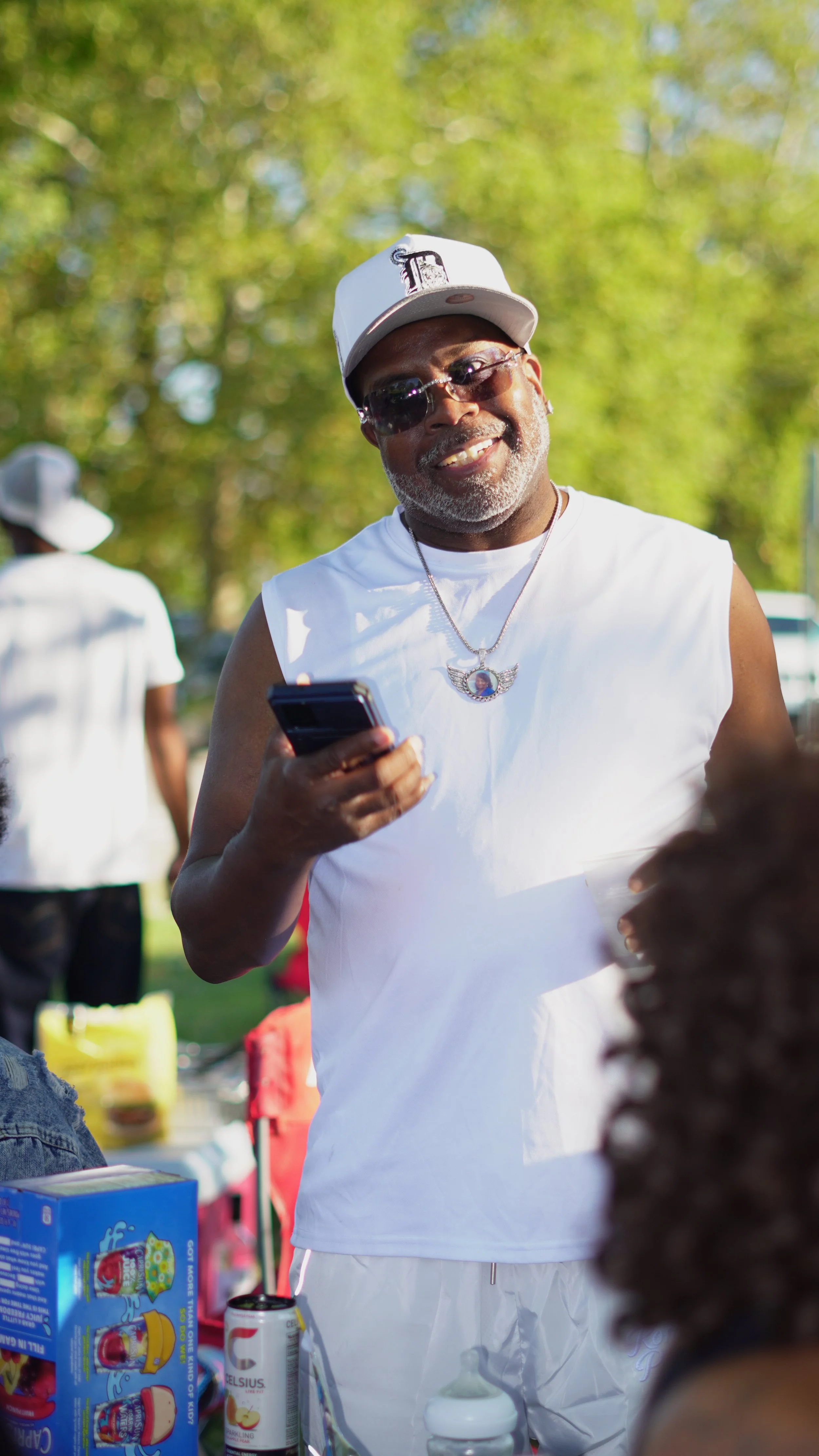 A smiling African American man wearing sunglasses, a white cap, sleeveless white shirt, and a chain with a pendant, holding a smartphone outdoors during daytime.