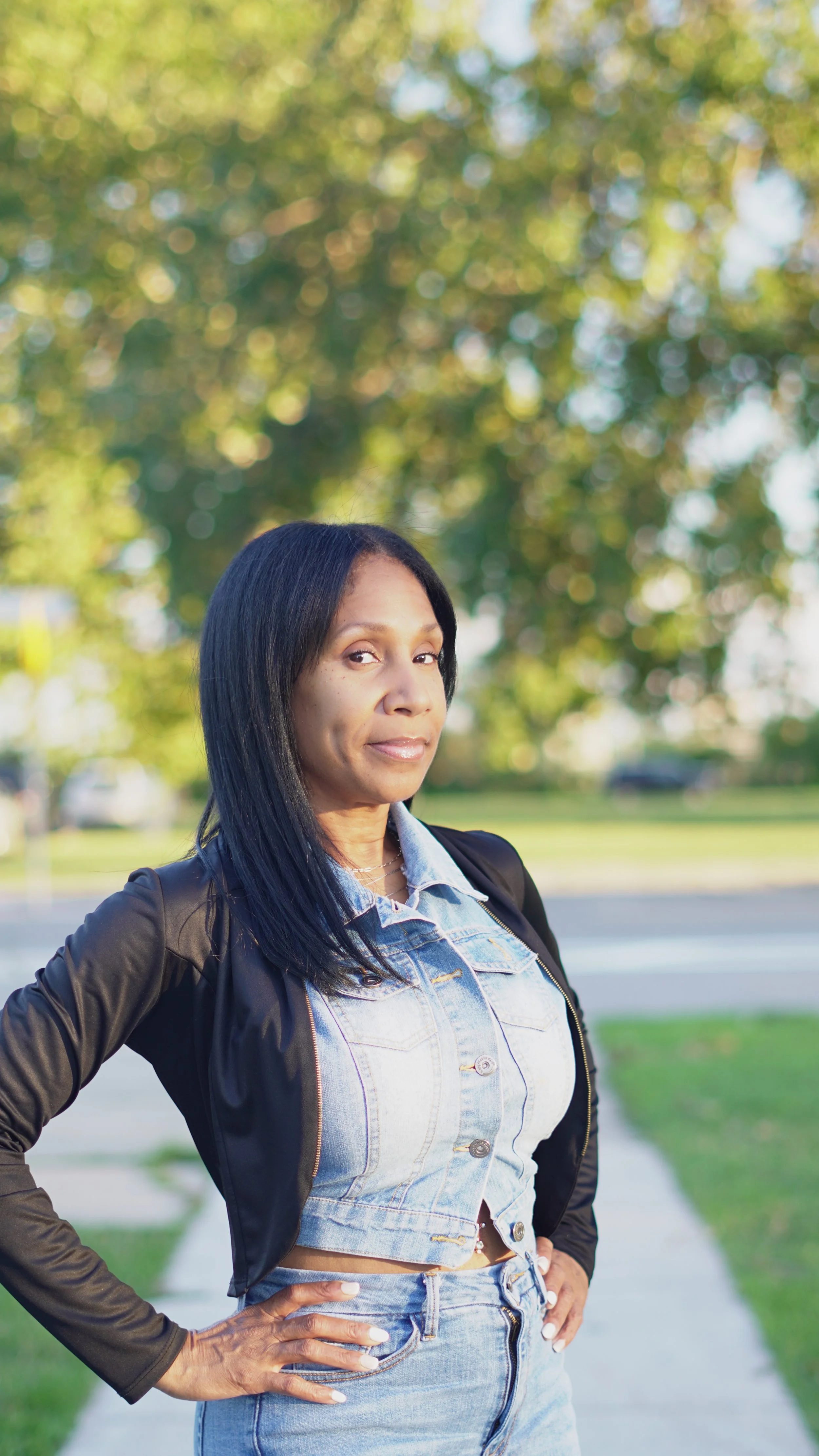 A woman with long black hair, wearing a denim jacket, crop top, and jeans, standing outdoors with her hand on her hip, in front of a background of green trees and a sidewalk, during daytime.