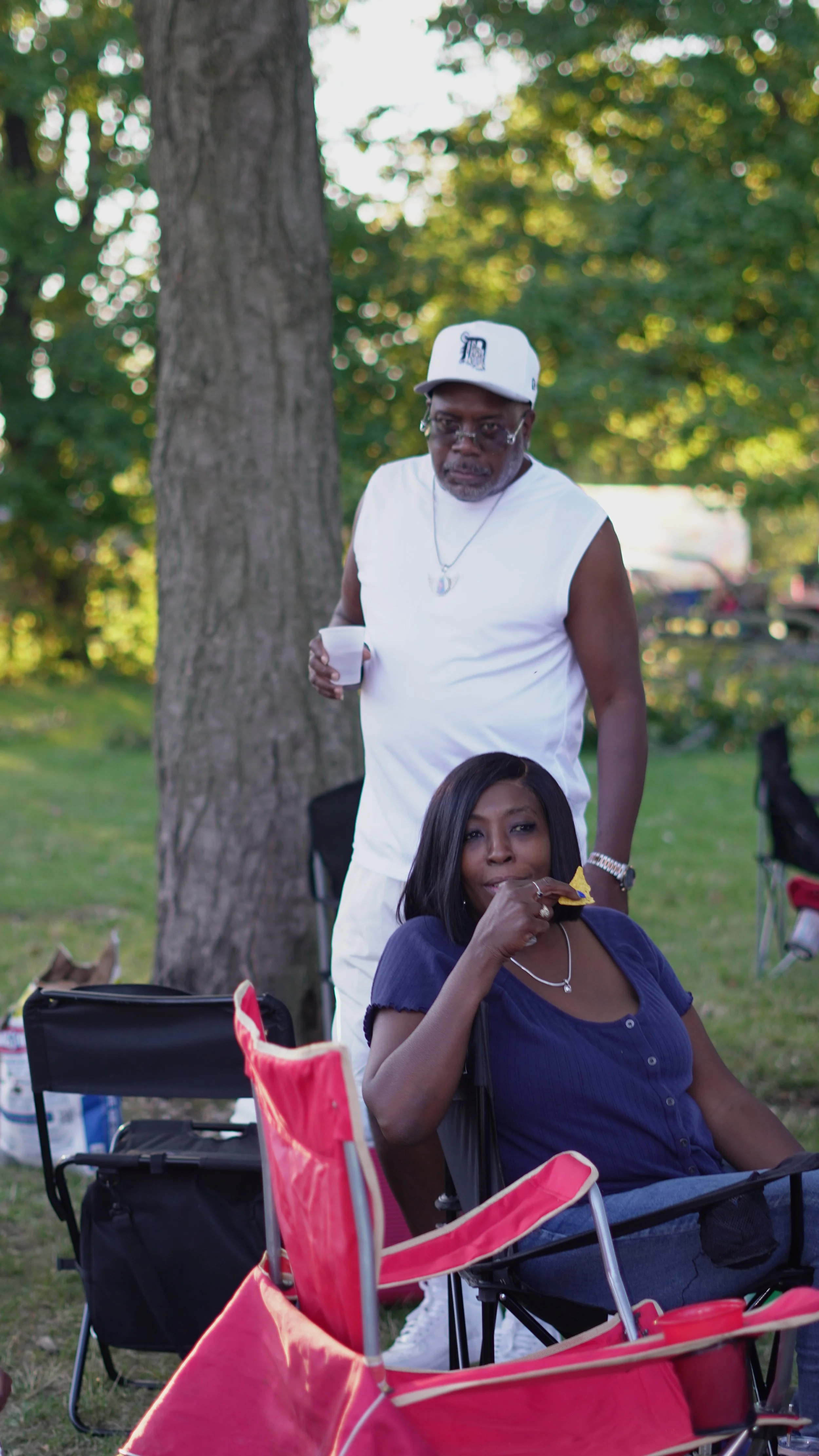 Two people at an outdoor gathering near a large tree. One man stands with a drink in his hand, wearing a white sleeveless shirt, white cap, and glasses. A woman sits in a folding chair, eating a snack, wearing a navy blue shirt, necklace, and has long dark hair. There are chairs, a bag, and other objects around them.